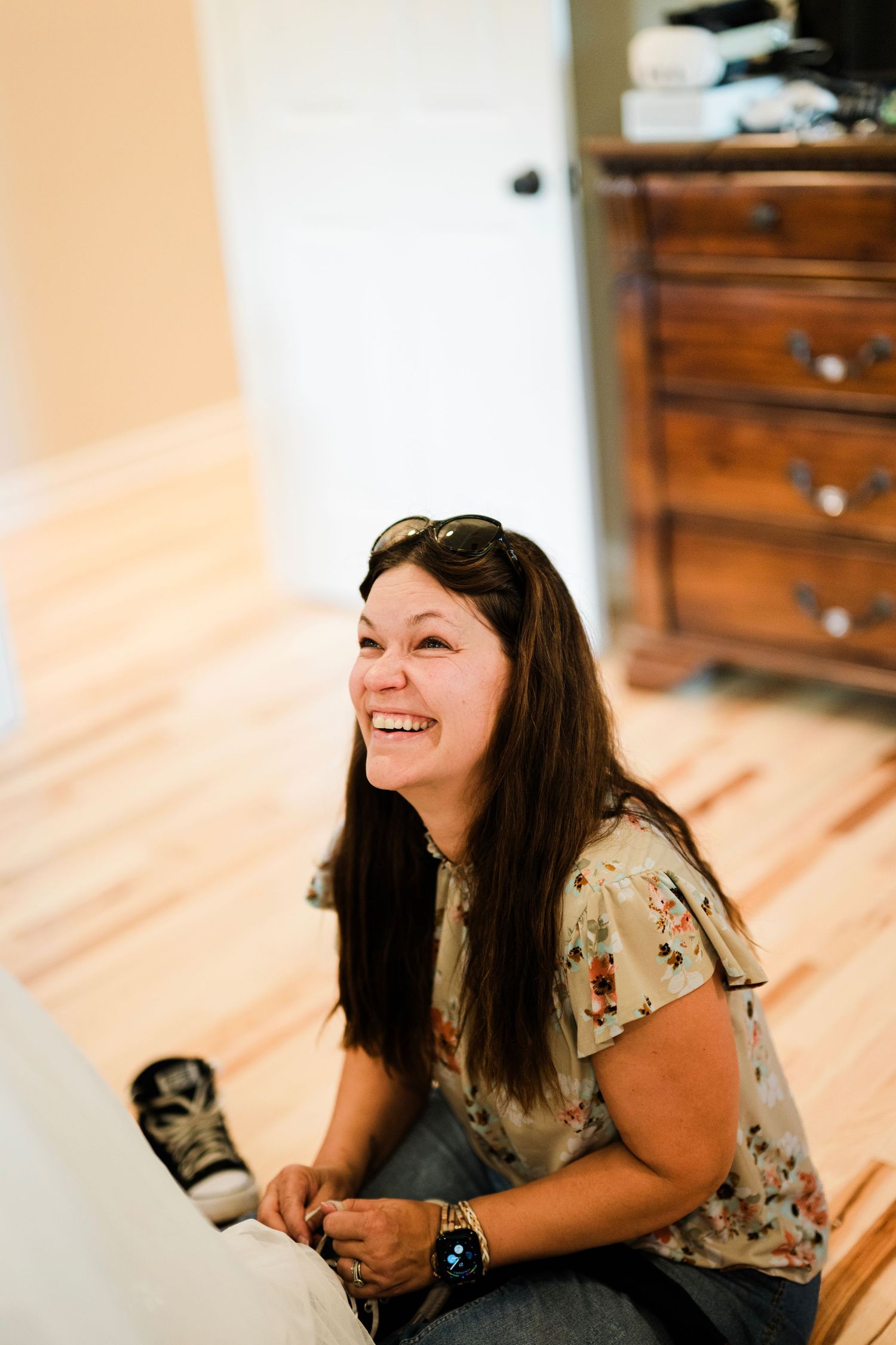 A laughing woman with sunglasses on her head sits on a hardwood floor in a floral blouse during a wedding preparation.