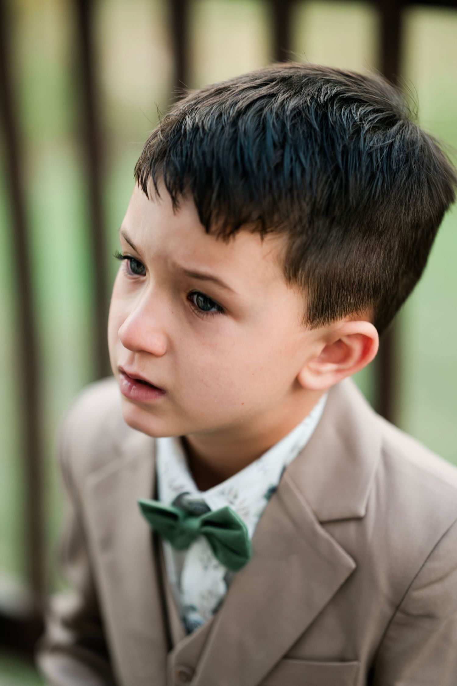 A young boy in a tan suit with a green bow tie gazes thoughtfully to the side at an outdoor wedding.