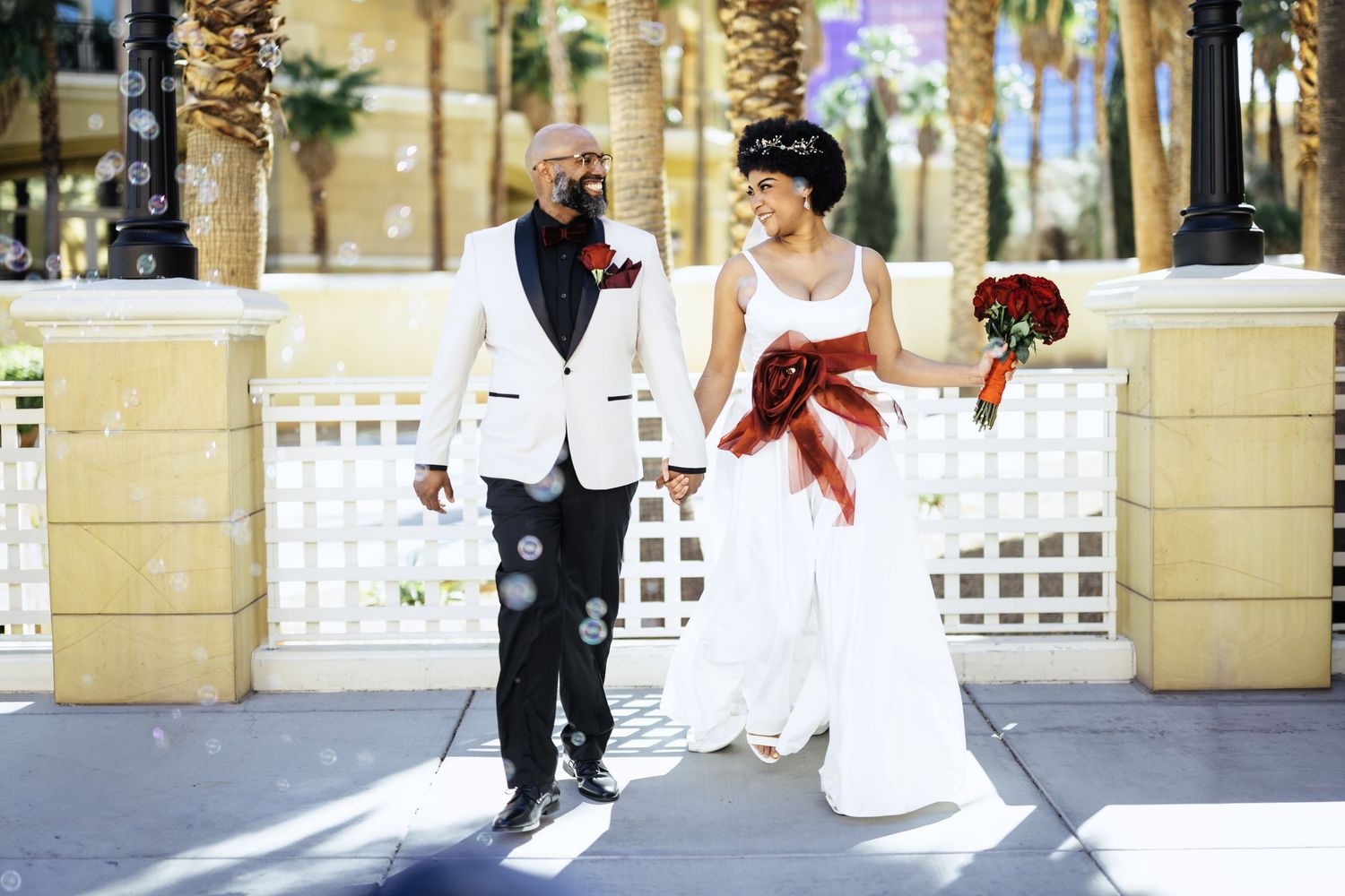 A stylish bride and groom walk hand in hand past palm trees, the groom in a white tuxedo with red accents.