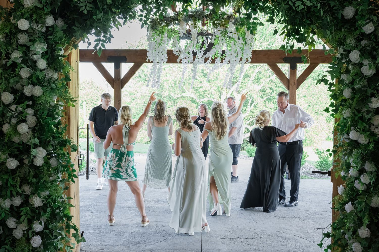 Wedding guests celebrate under floral arch with greenery and white roses at outdoor reception.