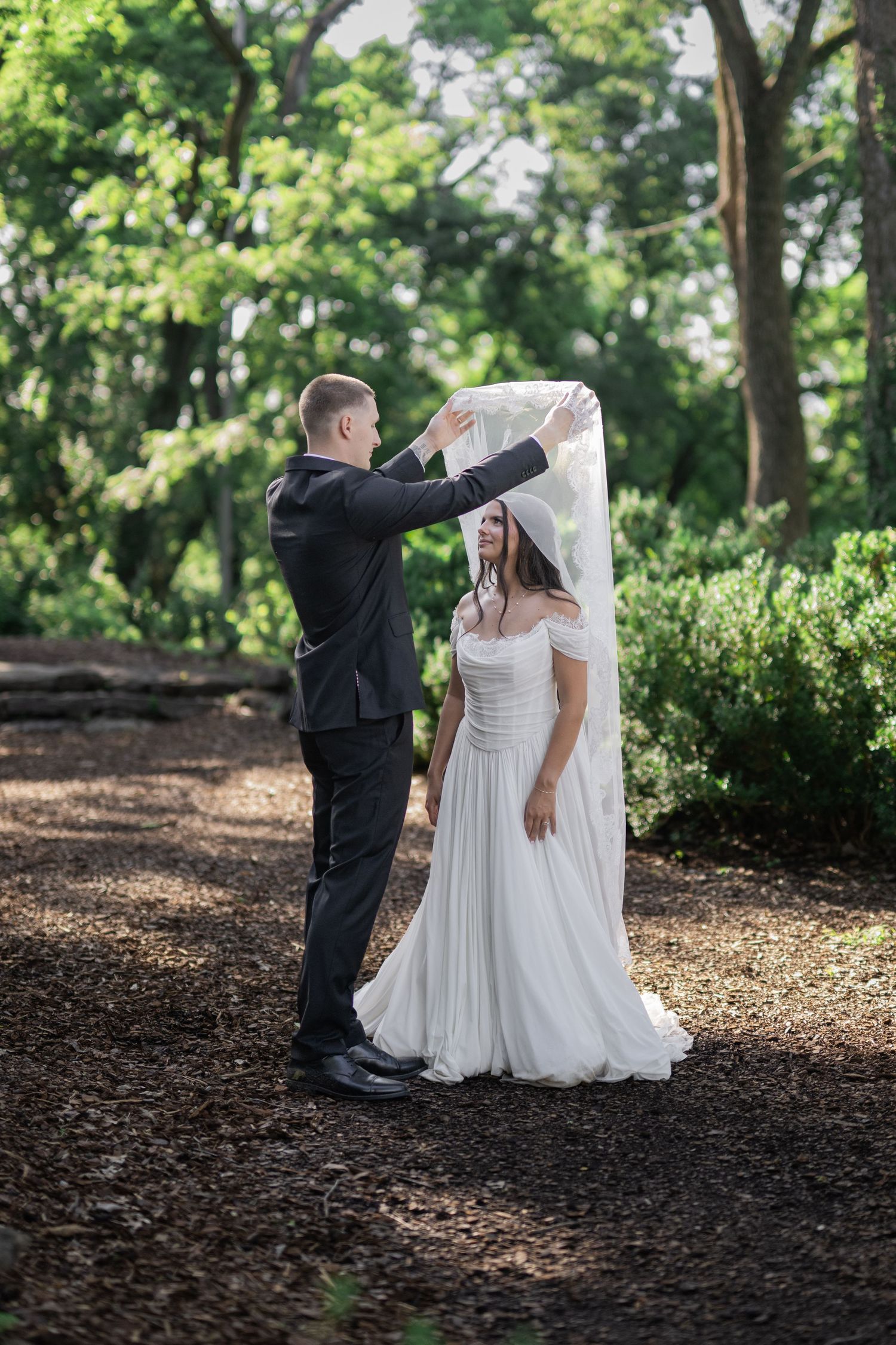 Groom lifting bride's veil in a lush green forest during an outdoor wedding ceremony.