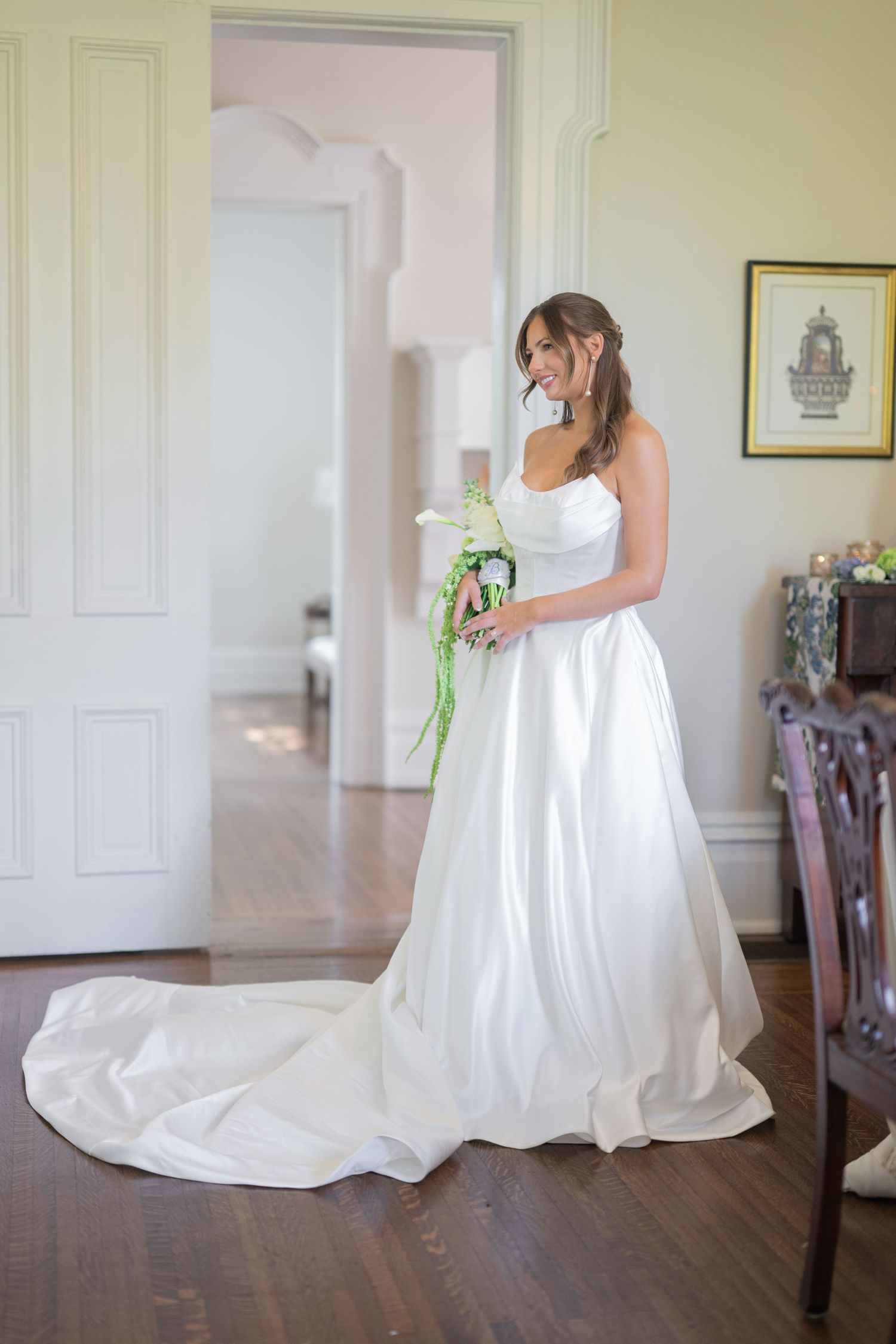 Bride in elegant white satin A-line gown with train holds floral bouquet in a bright, classic interior.