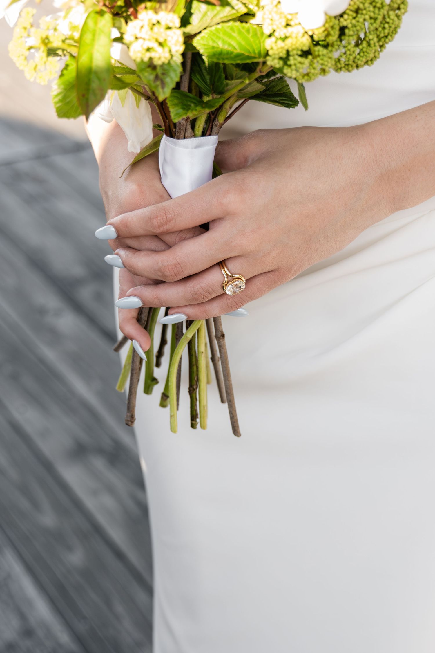 Bride holding a wedding bouquet of white and green flowers, wearing a gold engagement ring with light blue manicured nails.