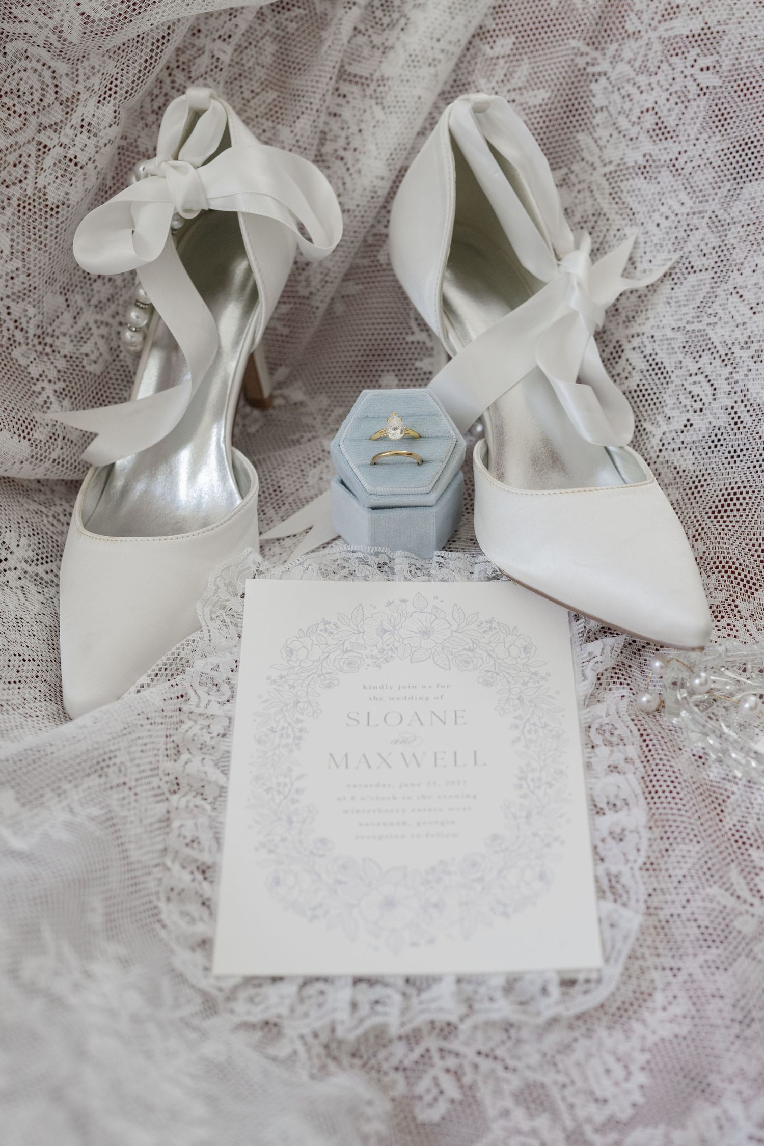 White bridal heels with ribbon ties beside a ring box and wedding invitation for Sloane and Maxwell on lace fabric.