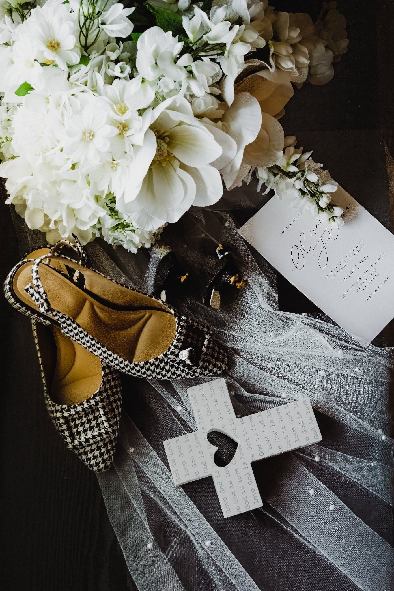 Flat lay of houndstooth bridal heels, white flowers, veil, and wedding invitation.