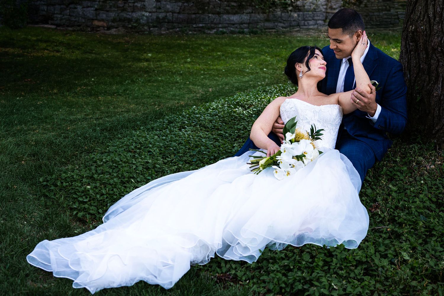 Bride in white gown and groom in navy suit sit together under a tree surrounded by lush green ivy.