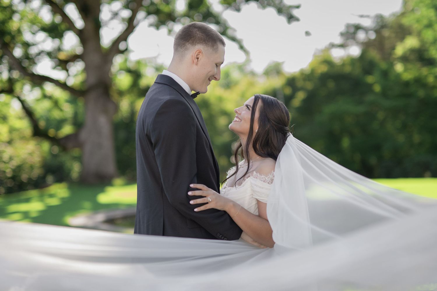 Bride and groom share a romantic moment outdoors, her veil flowing gracefully, surrounded by lush green trees.