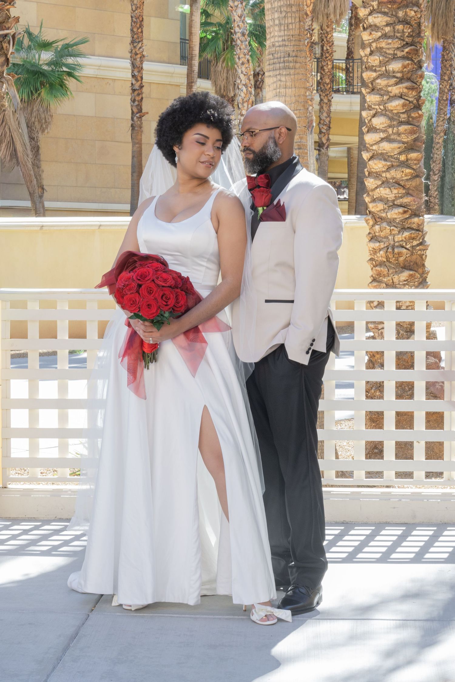 A bride in white gown holding red roses stands with her groom in a cream blazer outdoors near palm trees.