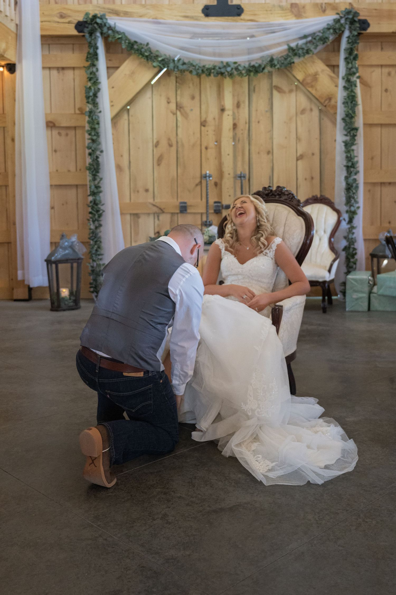 Groom kneeling before laughing bride in white gown inside rustic barn wedding venue with greenery garland backdrop.