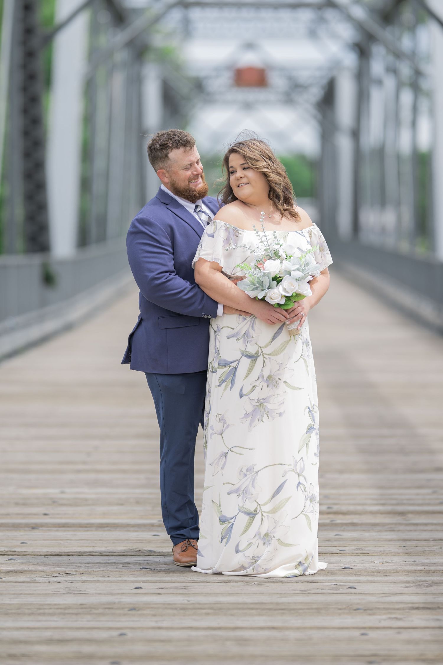 Bride and groom pose on a wooden bridge, she holds a floral bouquet in a patterned gown, he wears a navy suit.