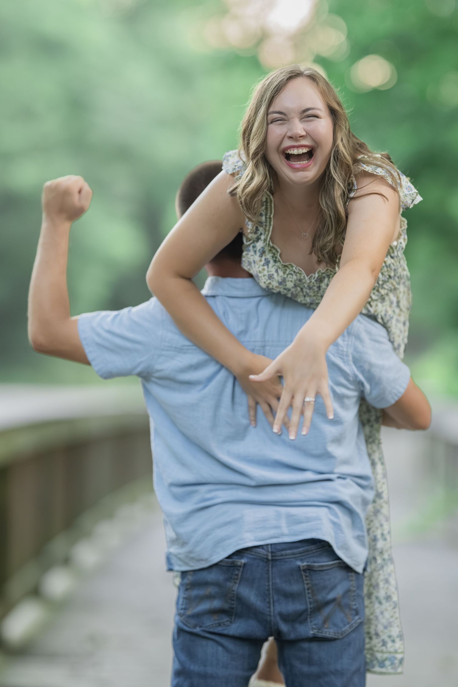 A joyful woman laughs while being carried piggyback by a man on a wooden bridge surrounded by green trees.