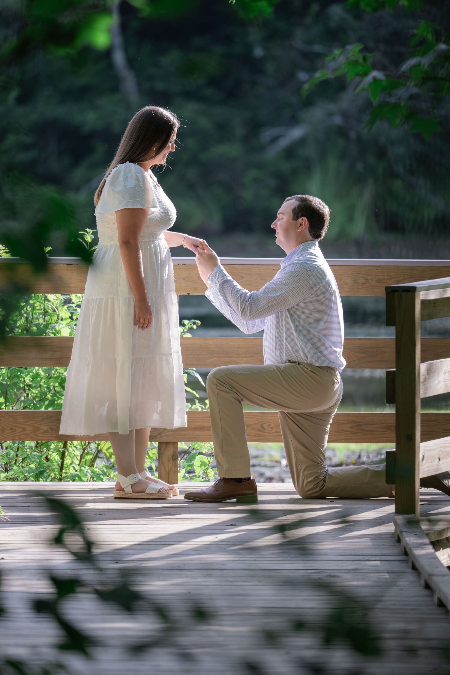 Man kneeling on one knee proposing to woman in white dress on wooden bridge surrounded by lush green trees.