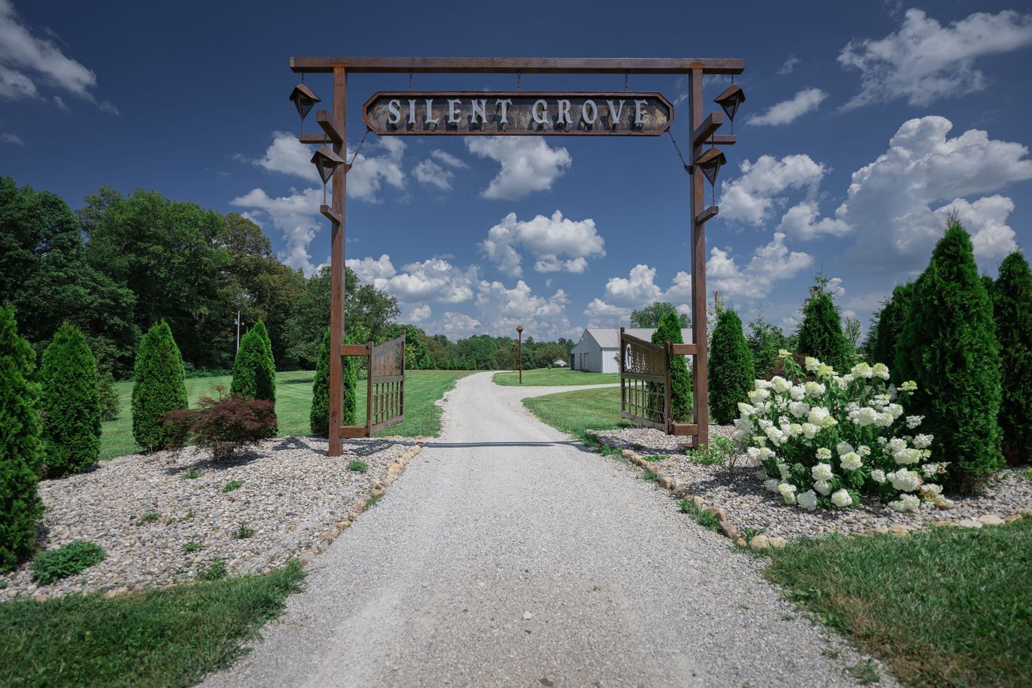 Rustic wooden entrance gate reading Silent Grove flanked by evergreen trees and white hydrangeas along a gravel driveway.