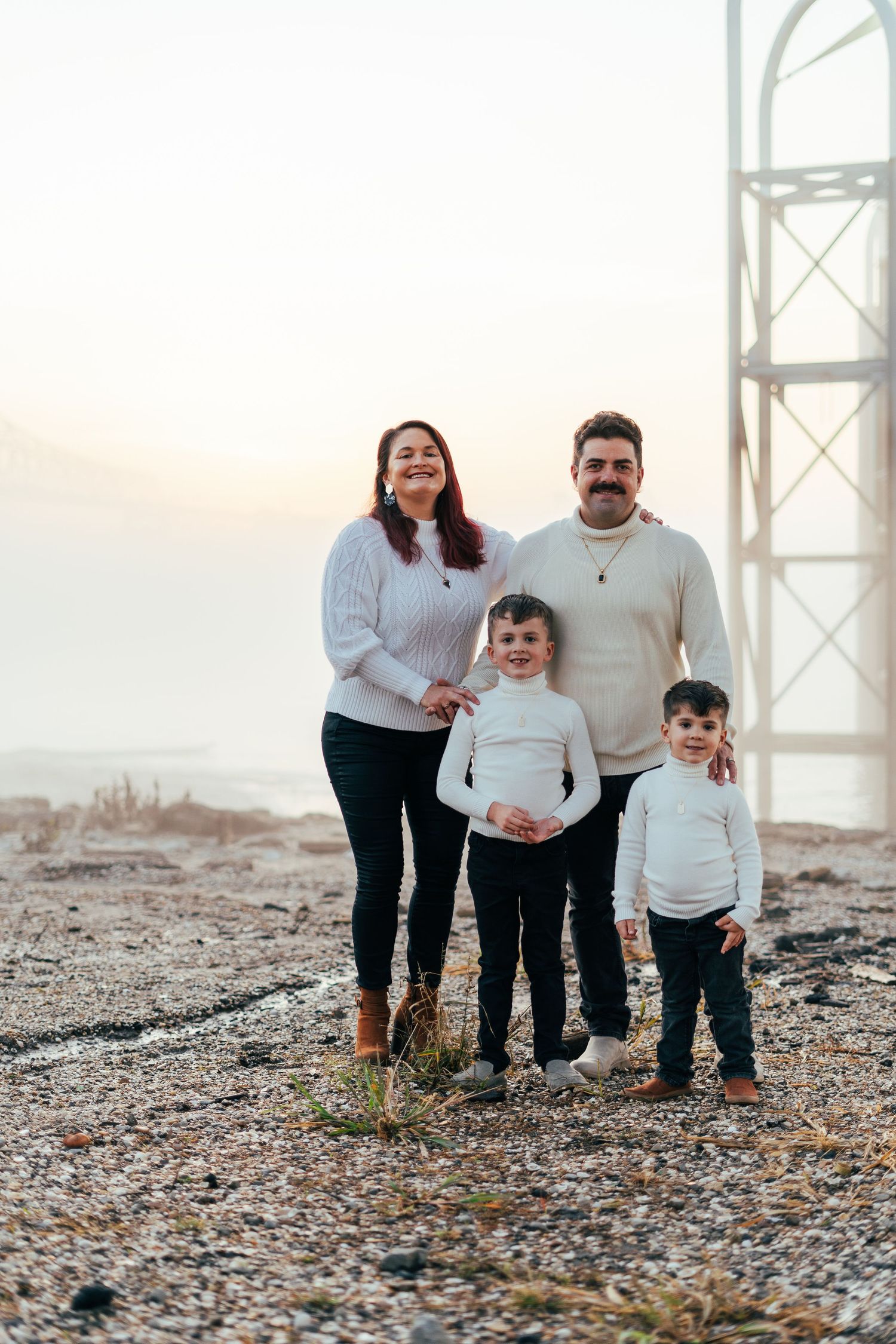 A family in coordinating white outfits poses together on a rocky beach at sunset.