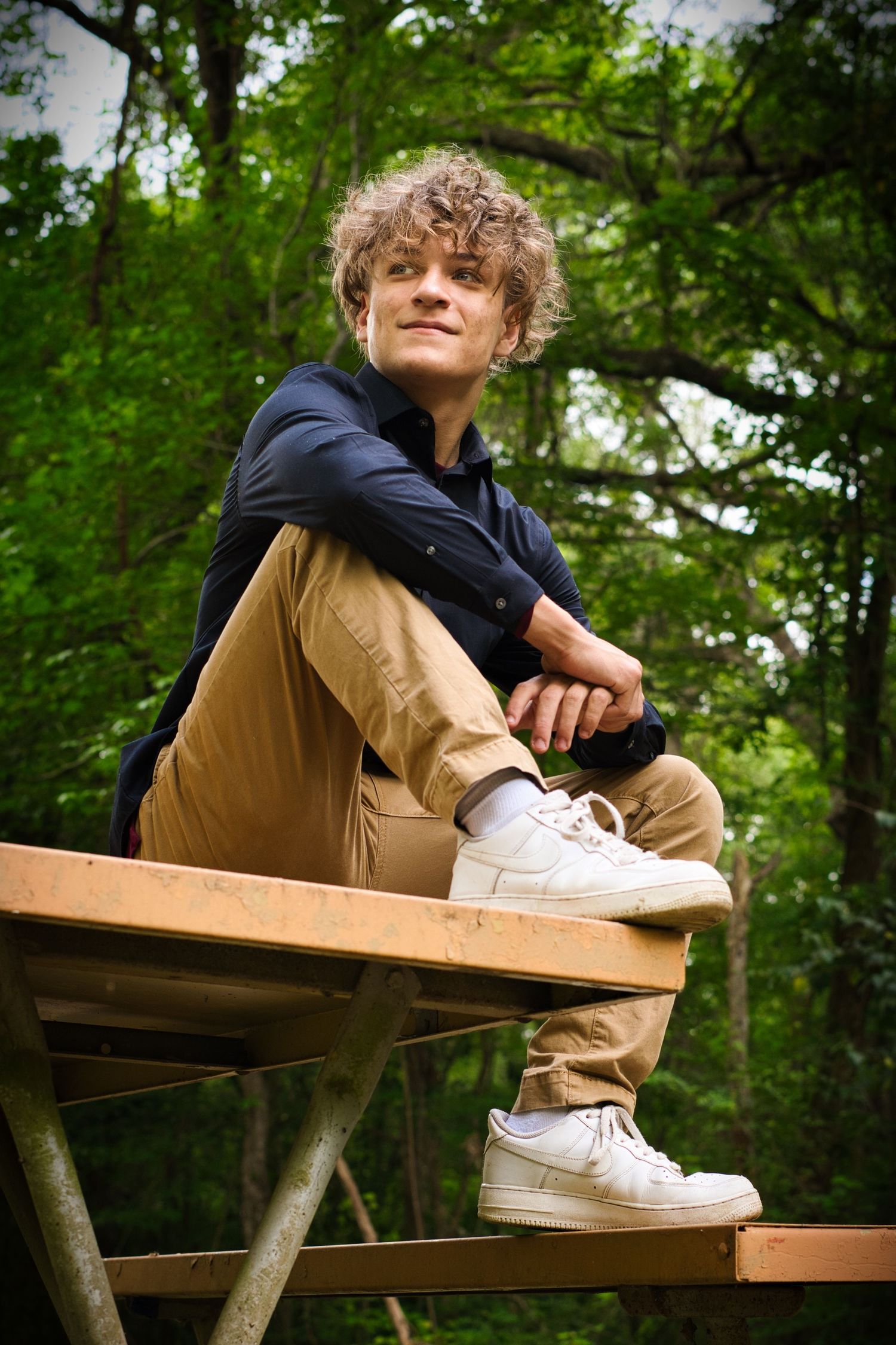 Person in navy hoodie and khakis sitting on wooden bench in wooded setting.