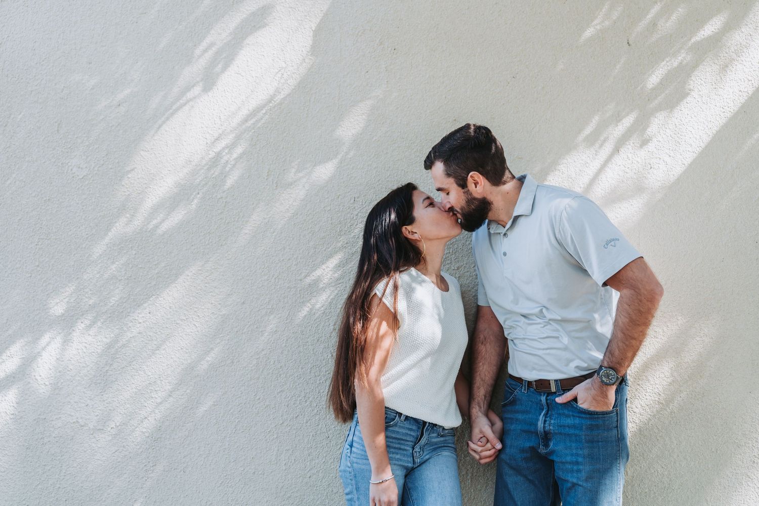 Couple shares intimate moments during a casual outdoor photo session against a white wall.
