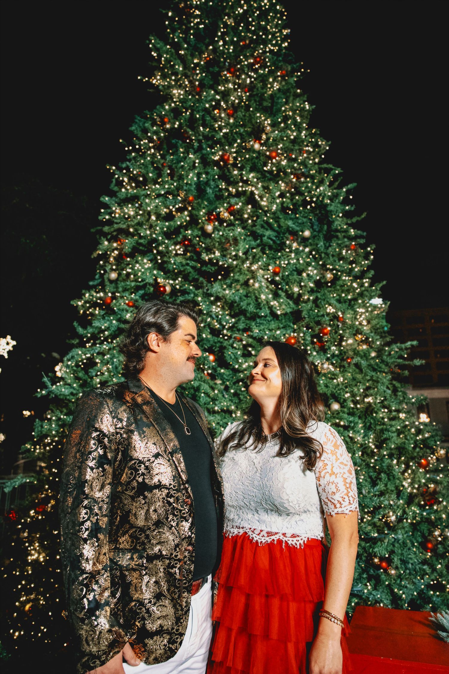 Romantic couple in formal attire posing by illuminated Christmas tree.