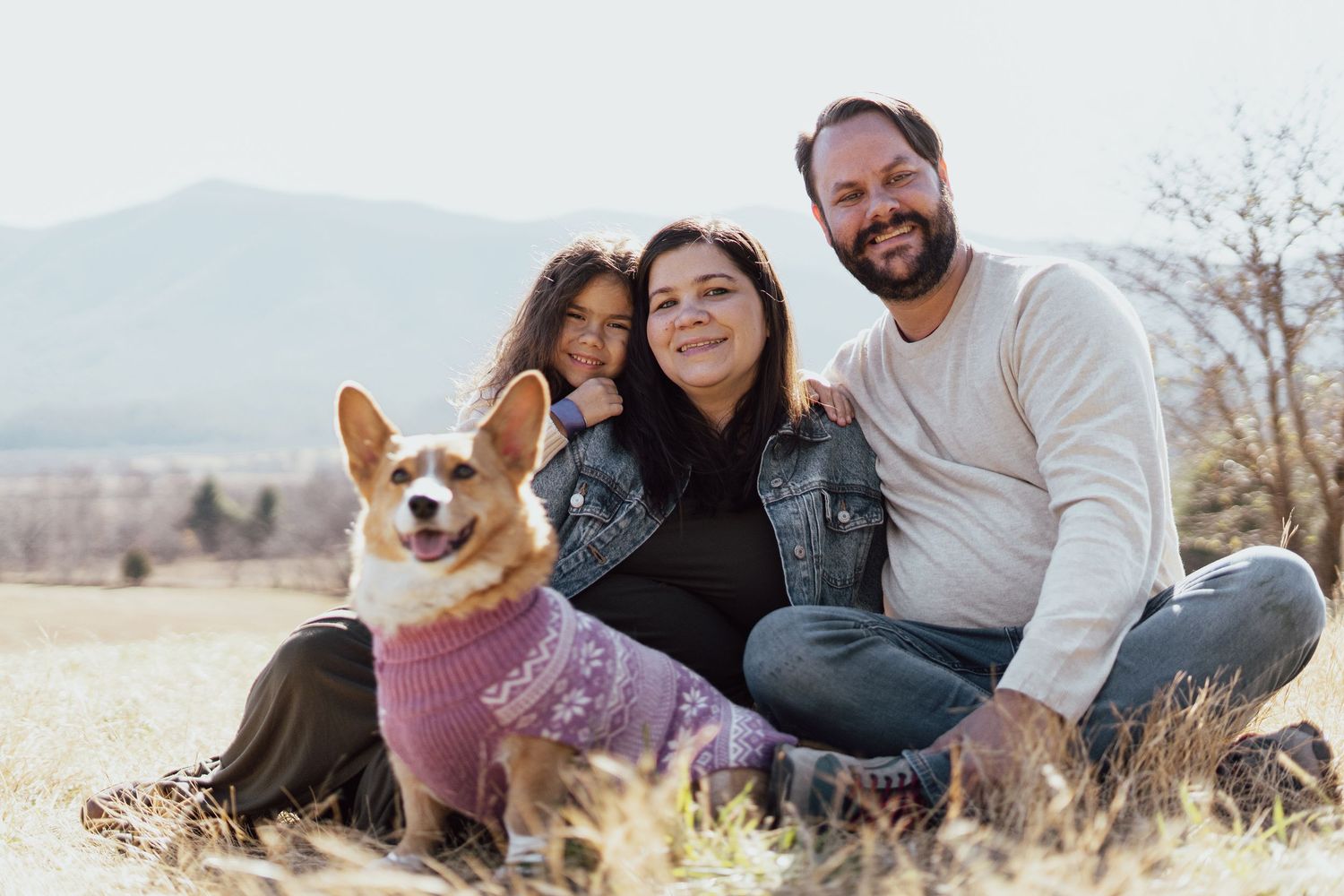 A couple and their Corgi dog pose together in a sunny field with mountains in the background.