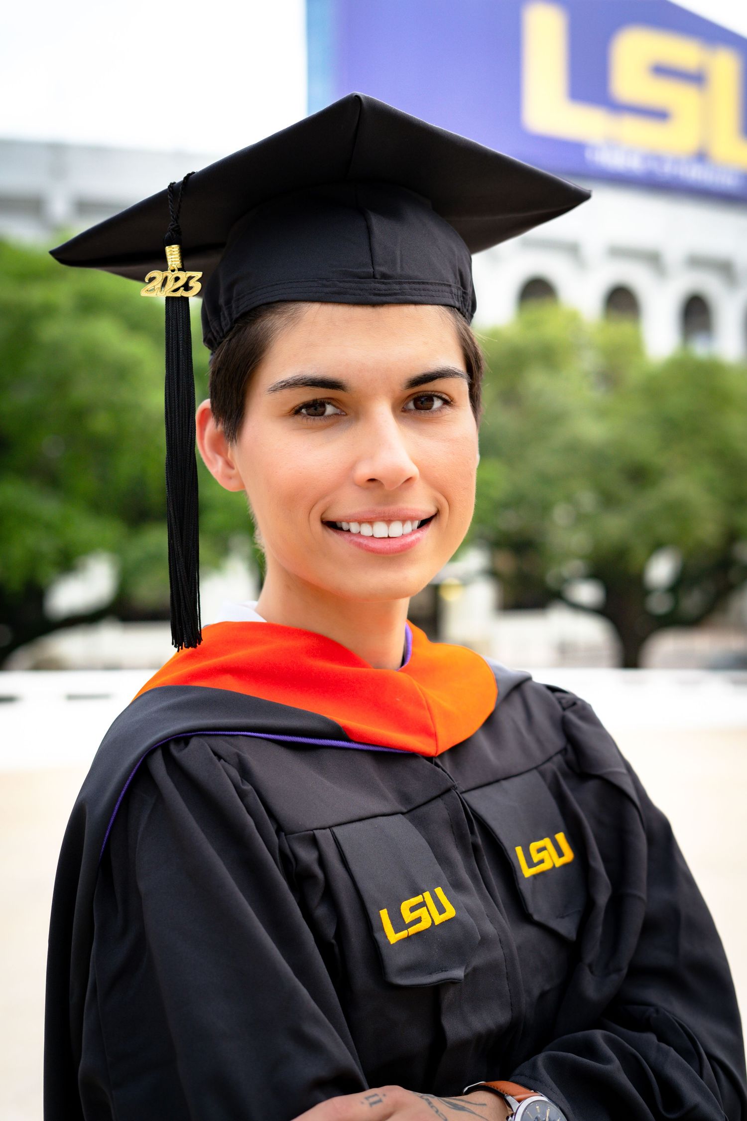 Graduate wearing black cap and gown with orange stole poses in front of LSU building.