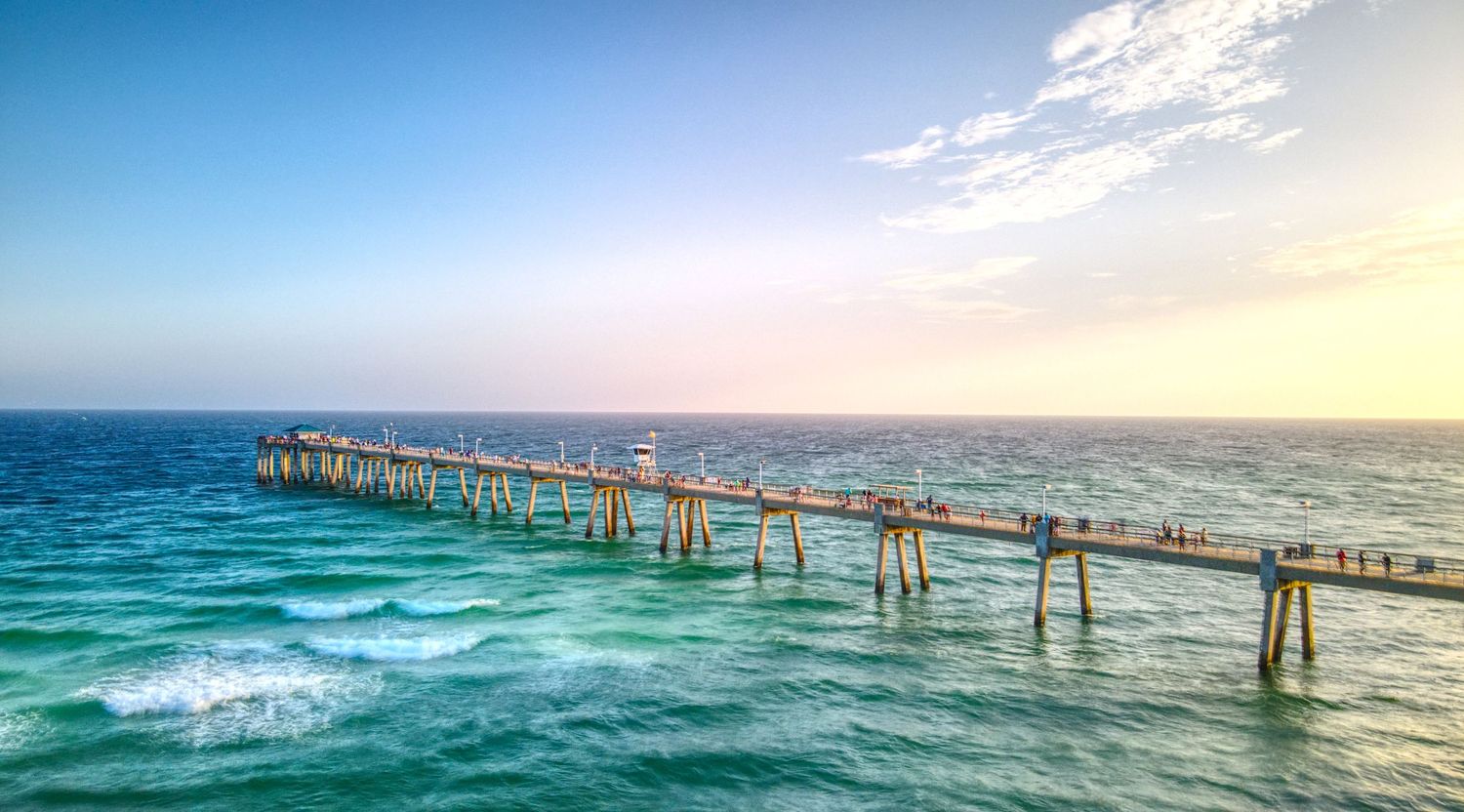 A wooden fishing pier extends over turquoise ocean waters under a pastel sunset sky.