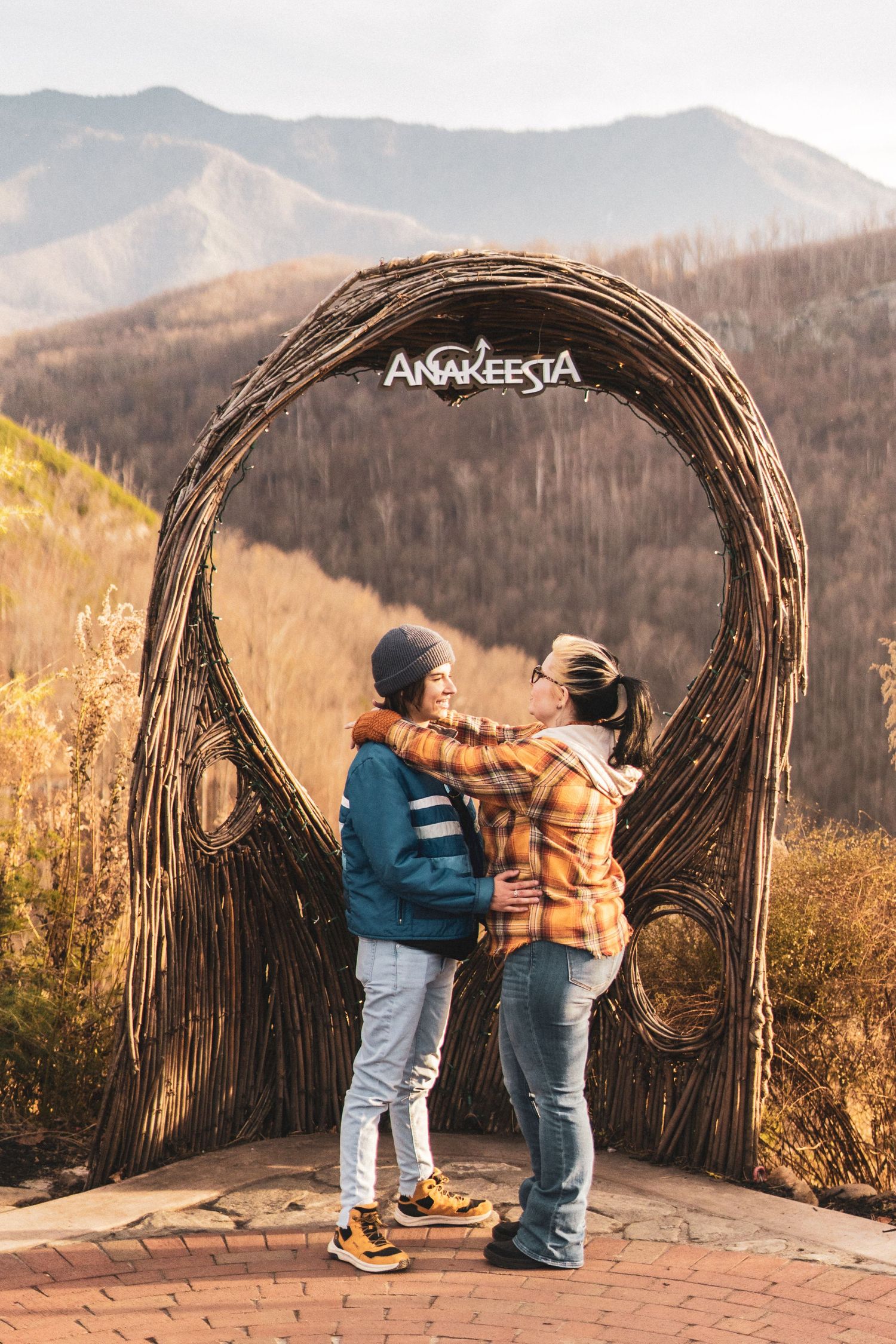 A couple shares a romantic moment under a rustic wooden arch overlooking mountain views at sunset.