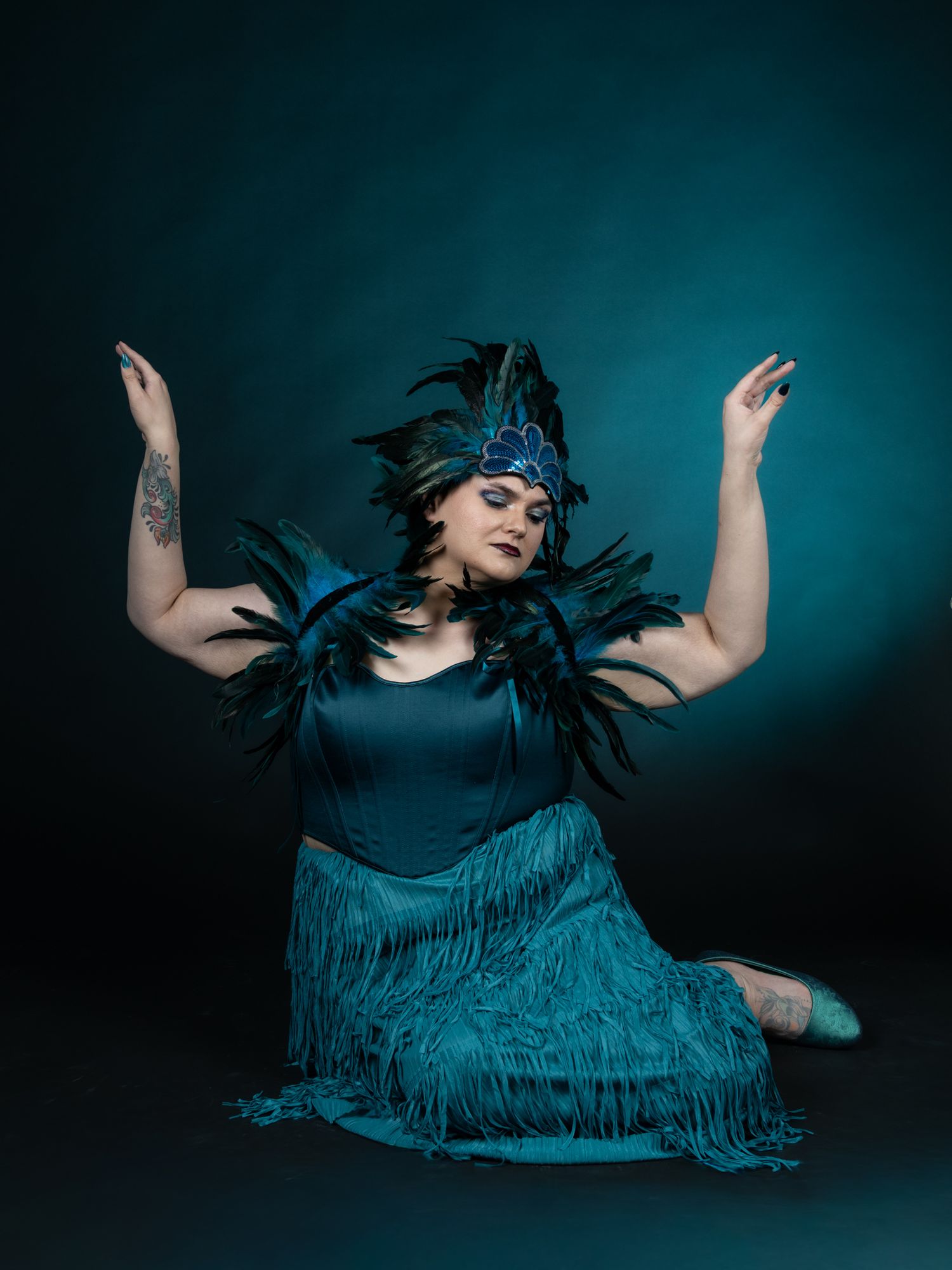 Model with teal tassle dress and feather headdress in front of black background with teal ombre