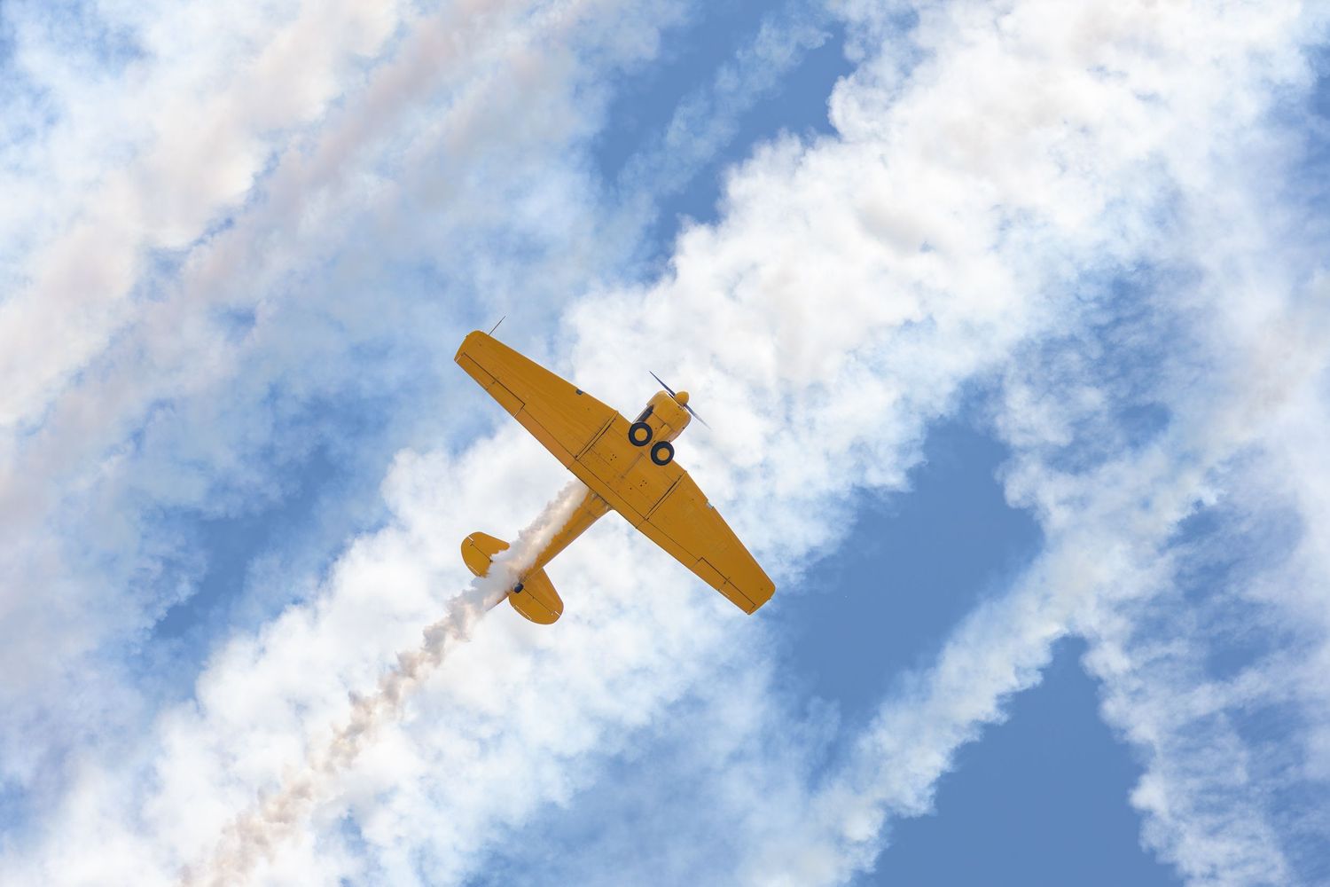 Bottom view of yellow plane flying with white smoke trails in the background