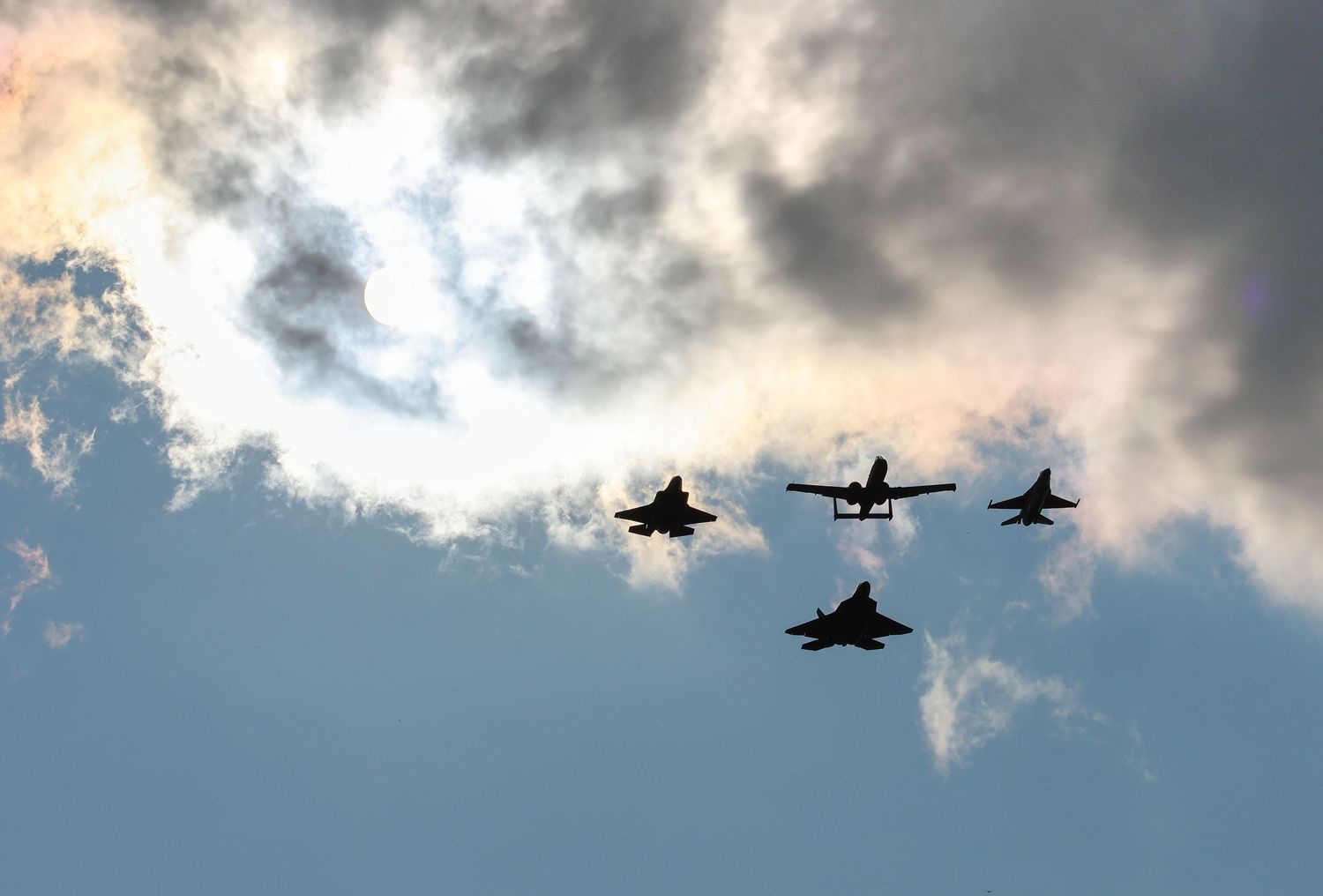 Silhouettes of F-35, A-10, F-22, and F-16 flying in formation in front of sun behind clouds