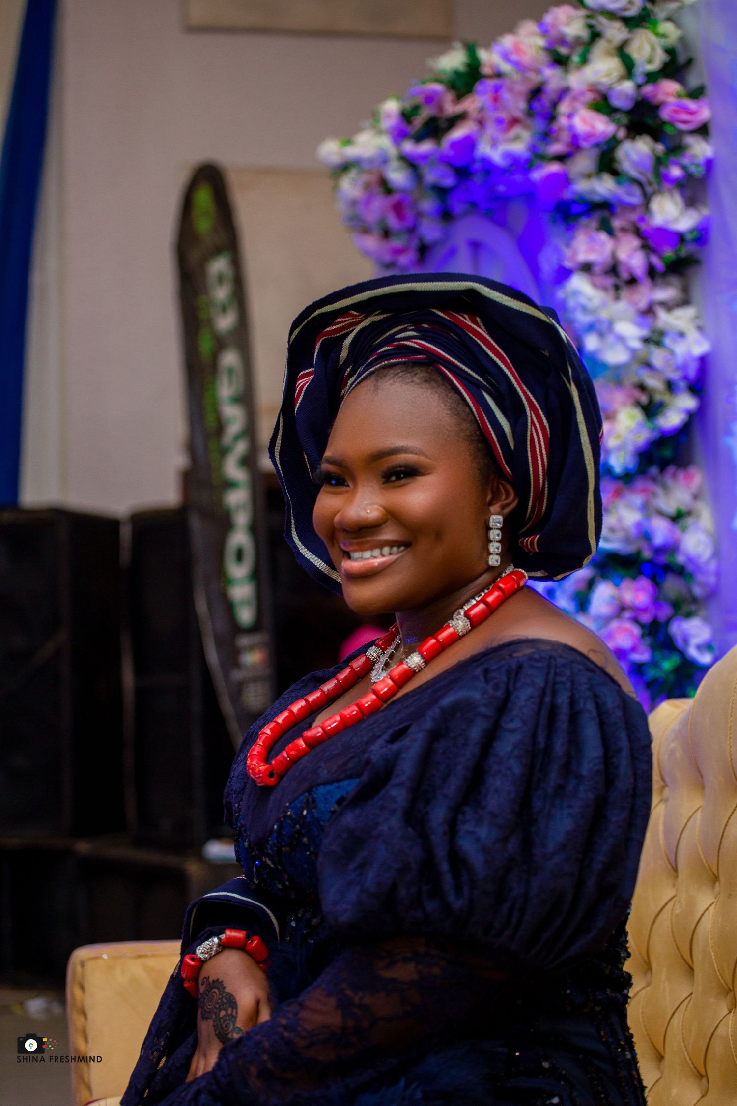 Radiant smile in navy traditional dress with coral beads against purple floral backdrop.