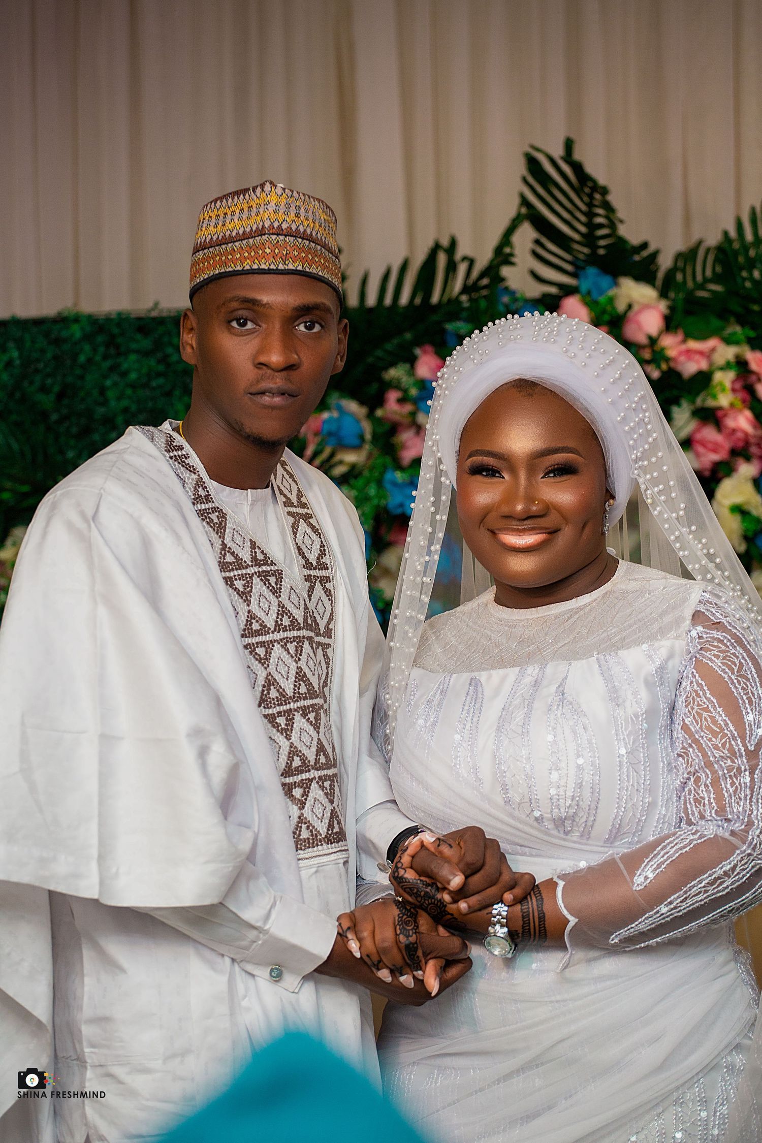 Wedding couple in traditional white attire and ceremonial headwear pose together against floral backdrop.