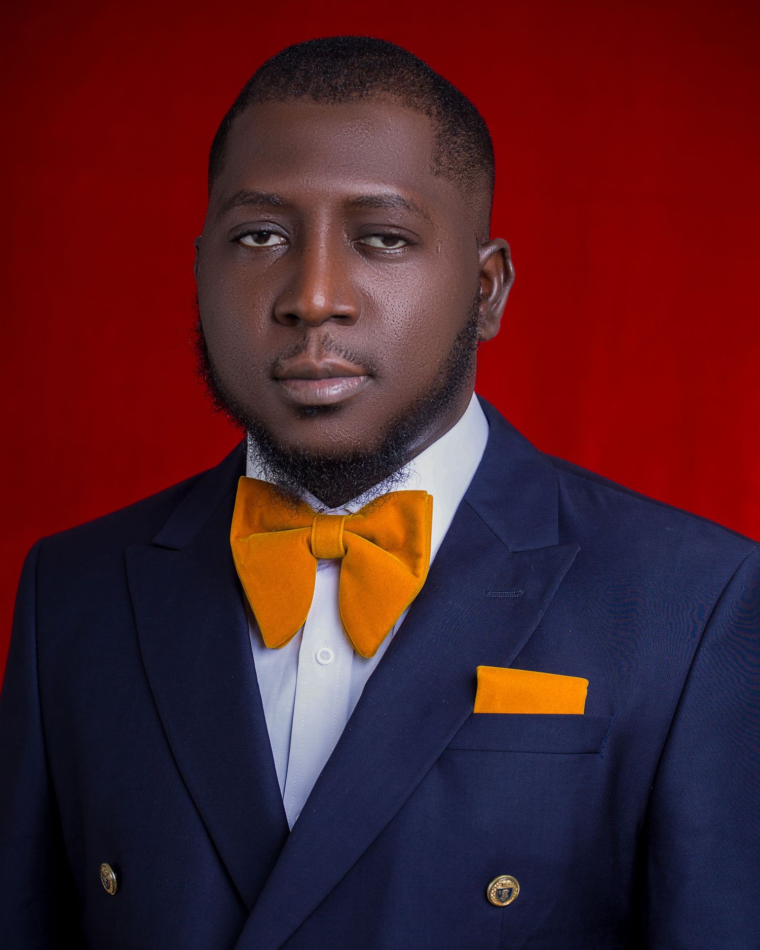 Professional studio portrait in navy suit with orange bow tie against red background.