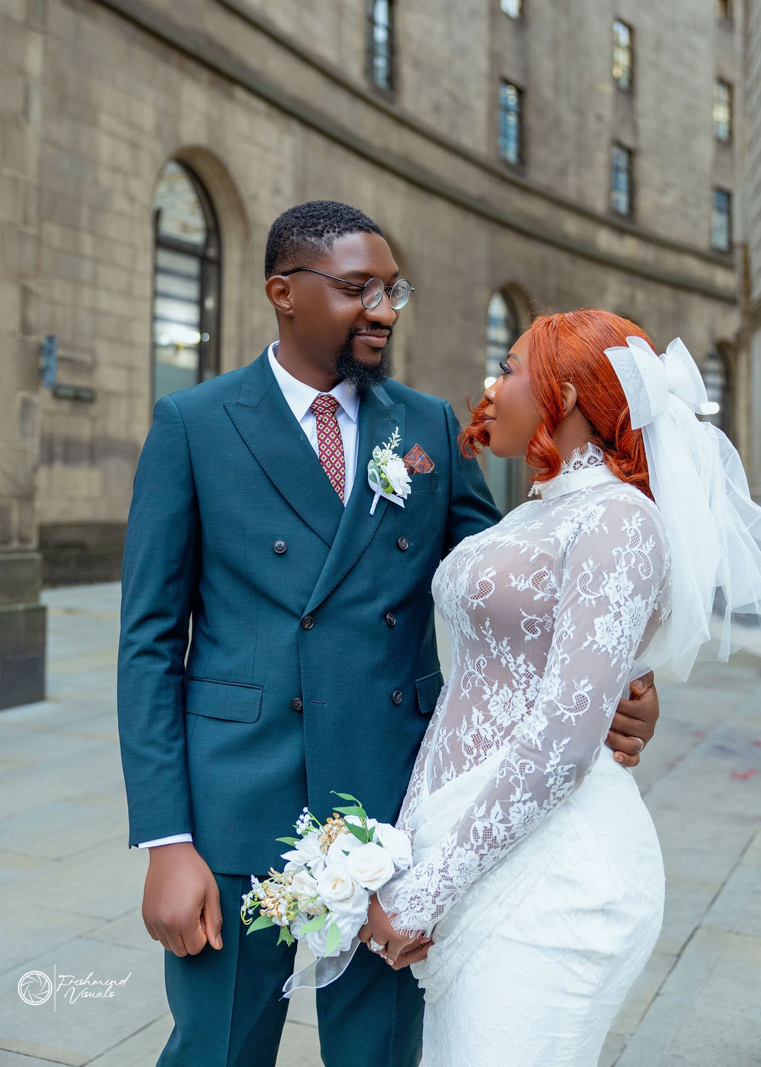 A couple in wedding attire shares a romantic moment outside a stone building, with the bride in lace and groom in navy suit.