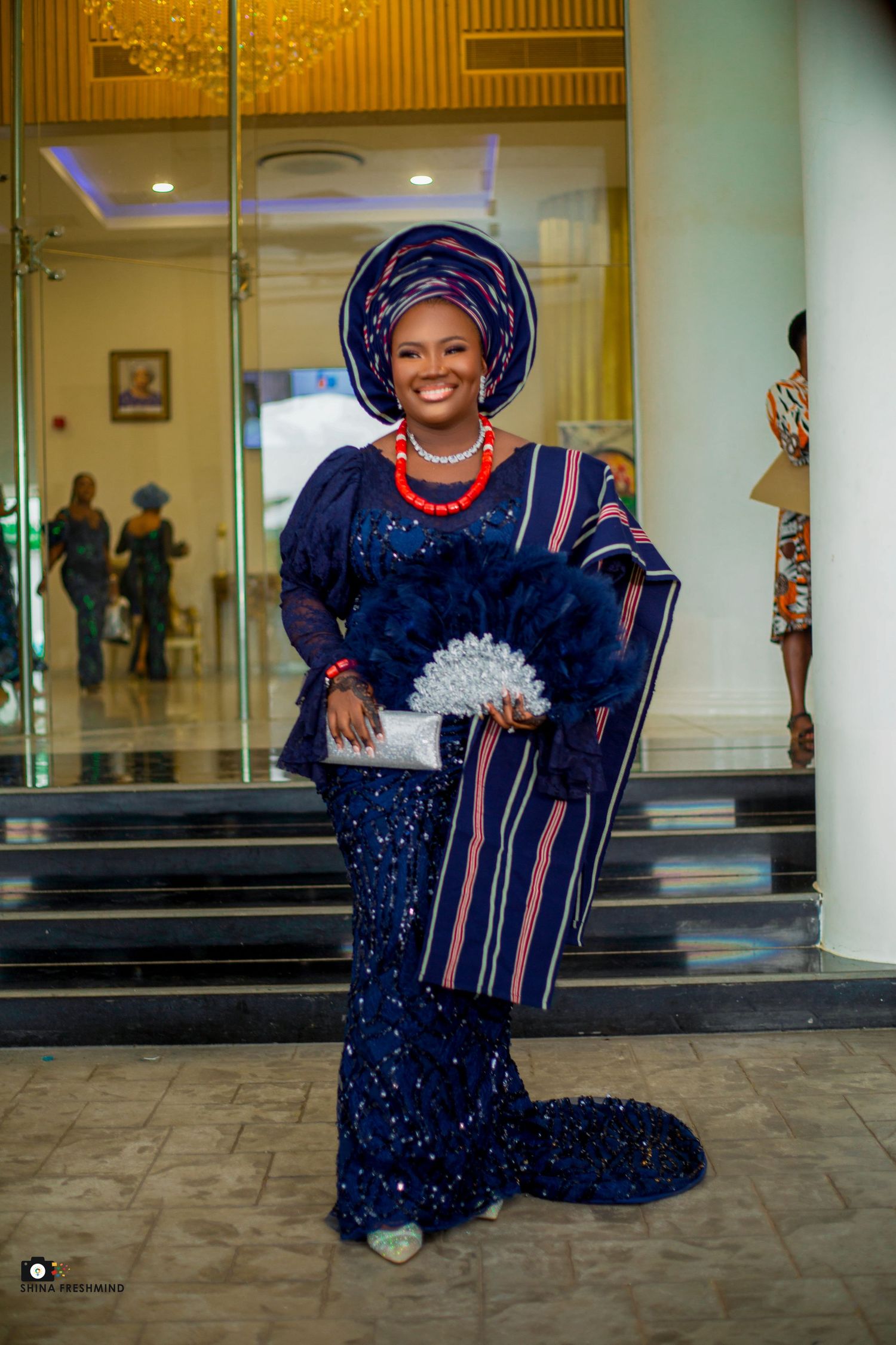 Elegant navy blue traditional outfit with matching gele and decorative hand fan at formal event.