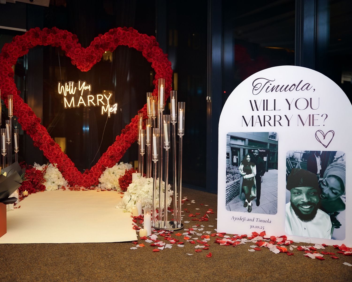 Romantic proposal setup with red rose heart arch and illuminated 'Will You Marry Me' sign.