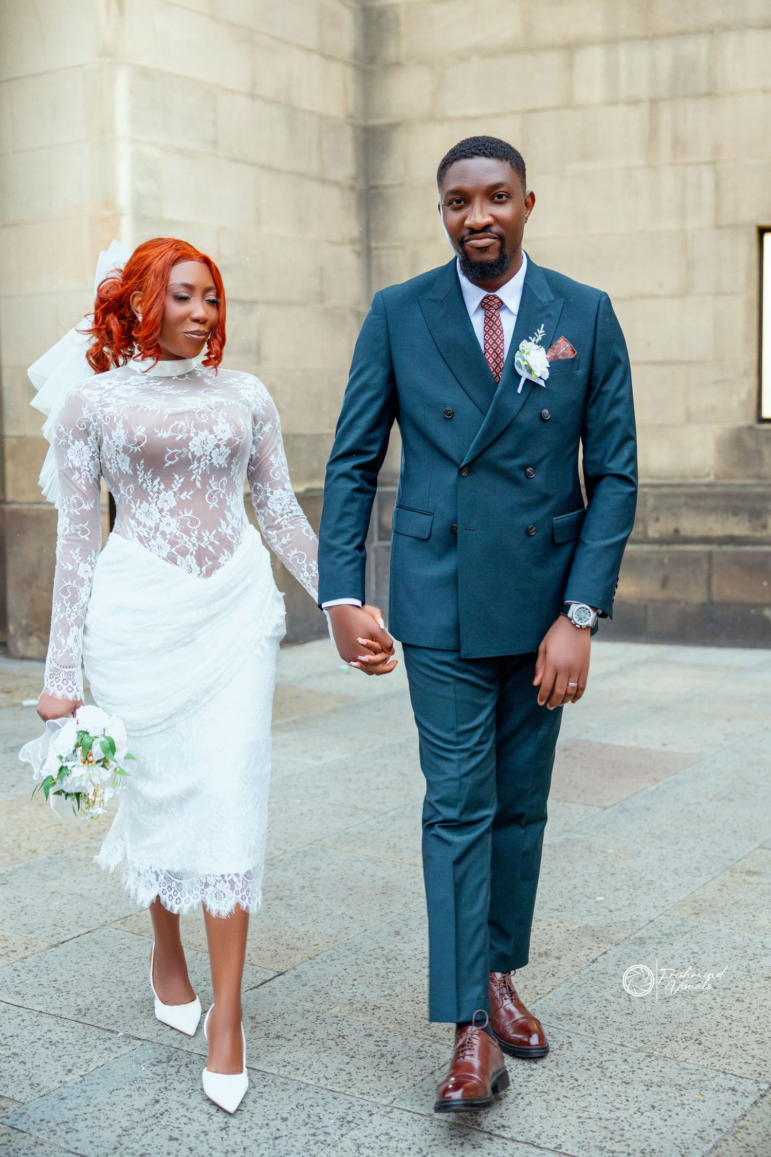 A couple in wedding attire walks together on concrete, with the bride wearing a short lace dress and groom in navy suit.