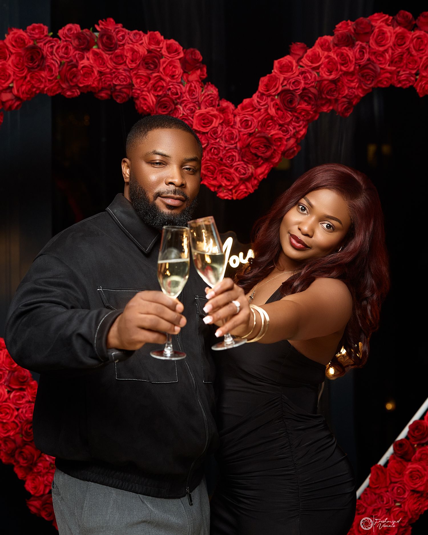 Couple toasting with champagne glasses under a red rose arch at an elegant event.