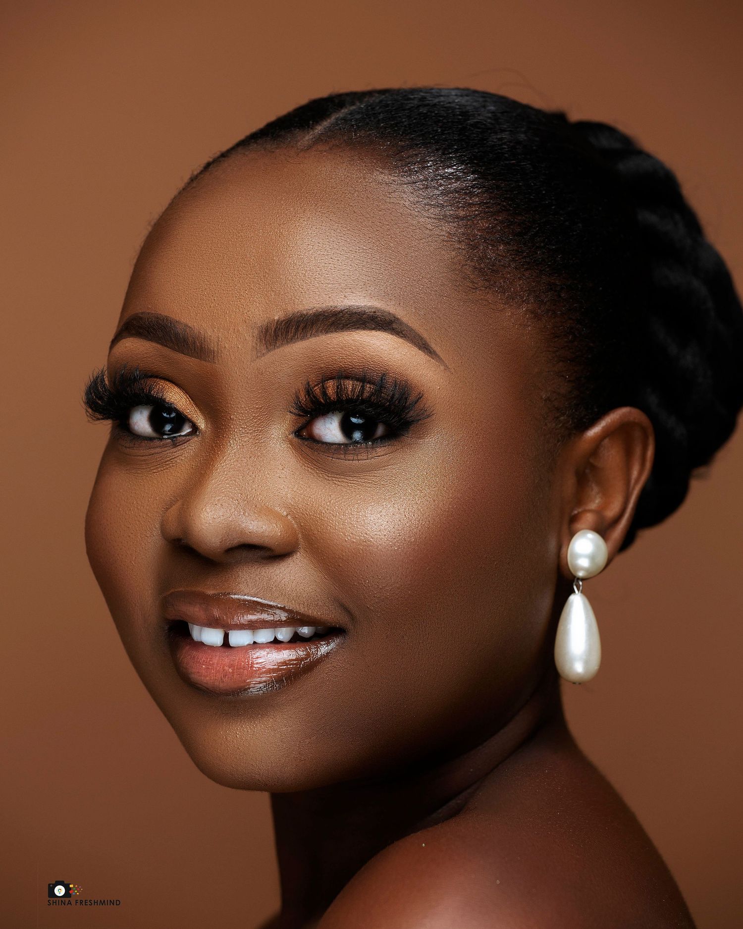 Glamour portrait with natural makeup, pearl earrings and updo hairstyle against warm brown background.