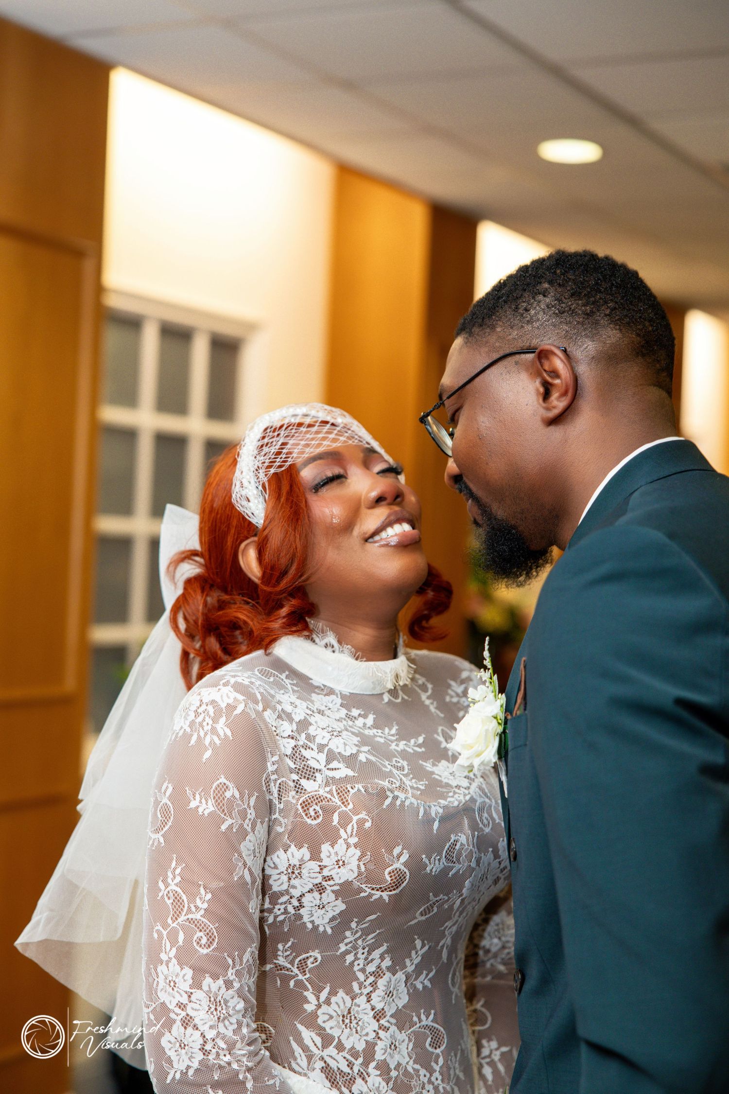 A couple shares an intimate moment at their wedding, with the bride in a lace dress and the groom in a dark green suit.