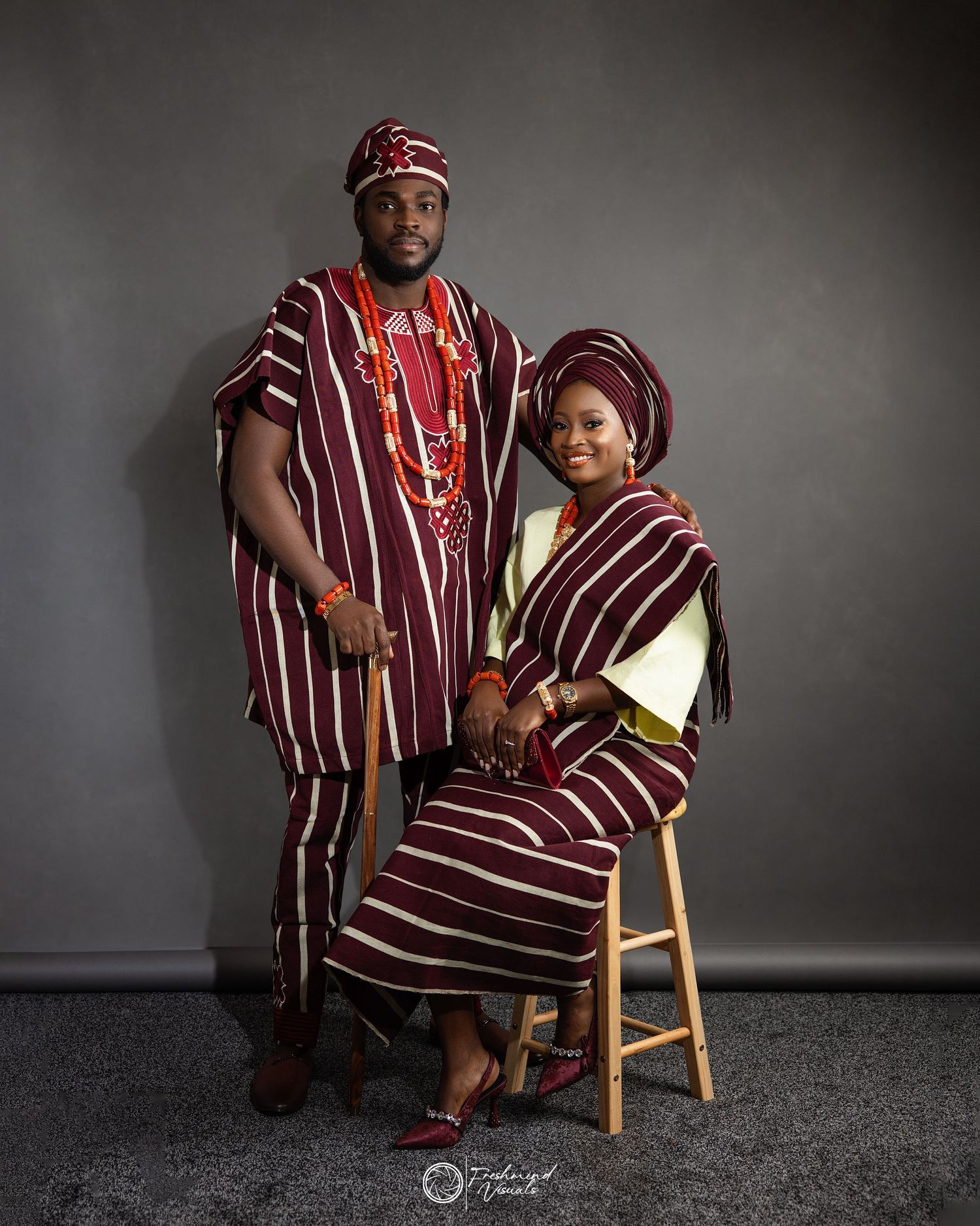A couple poses in matching burgundy striped traditional attire against a dark studio backdrop.