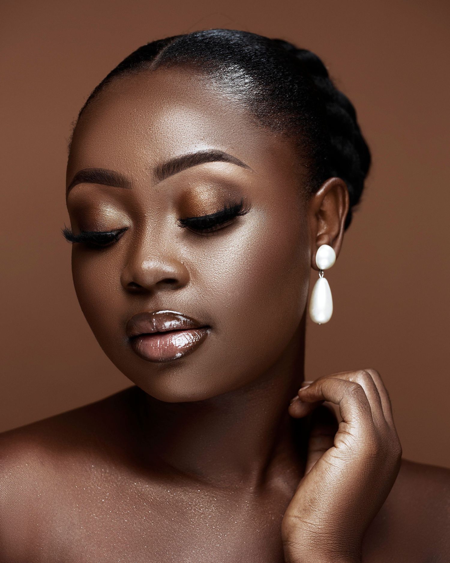 Moody beauty portrait with bronze tones, dramatic eye makeup and pearl earrings against brown backdrop.