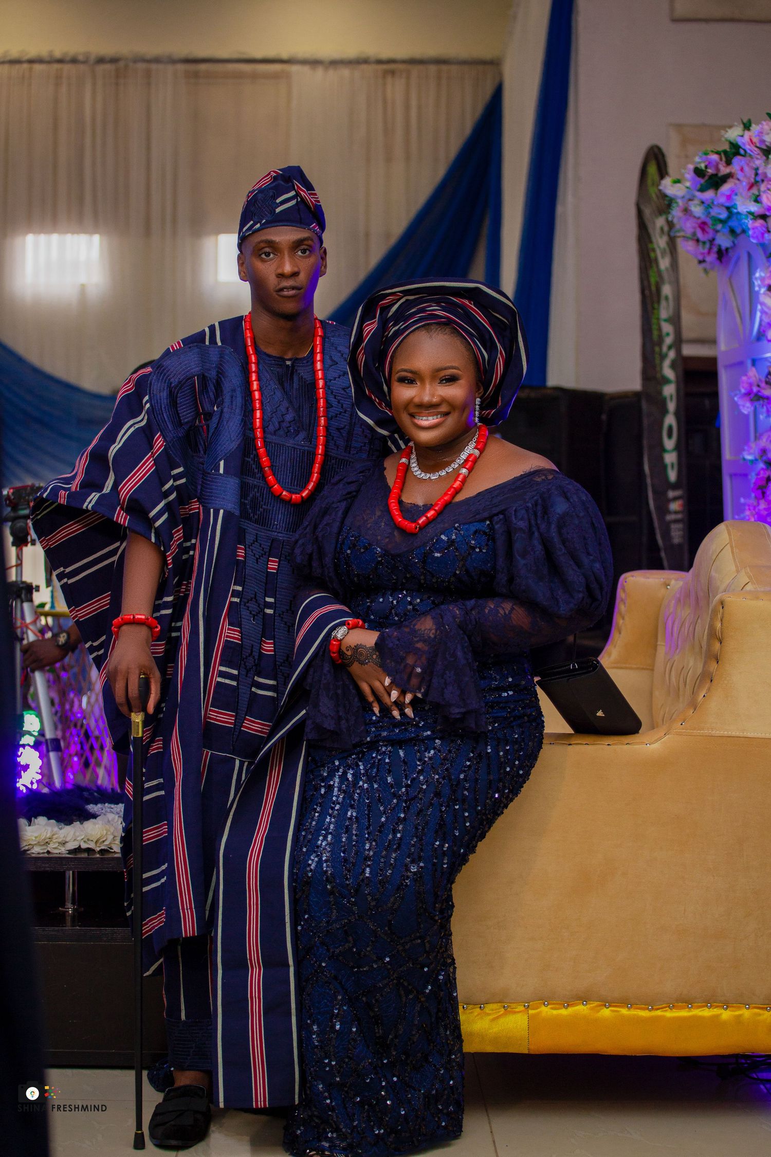 Couple in coordinated navy traditional attire posing against floral backdrop at celebration.