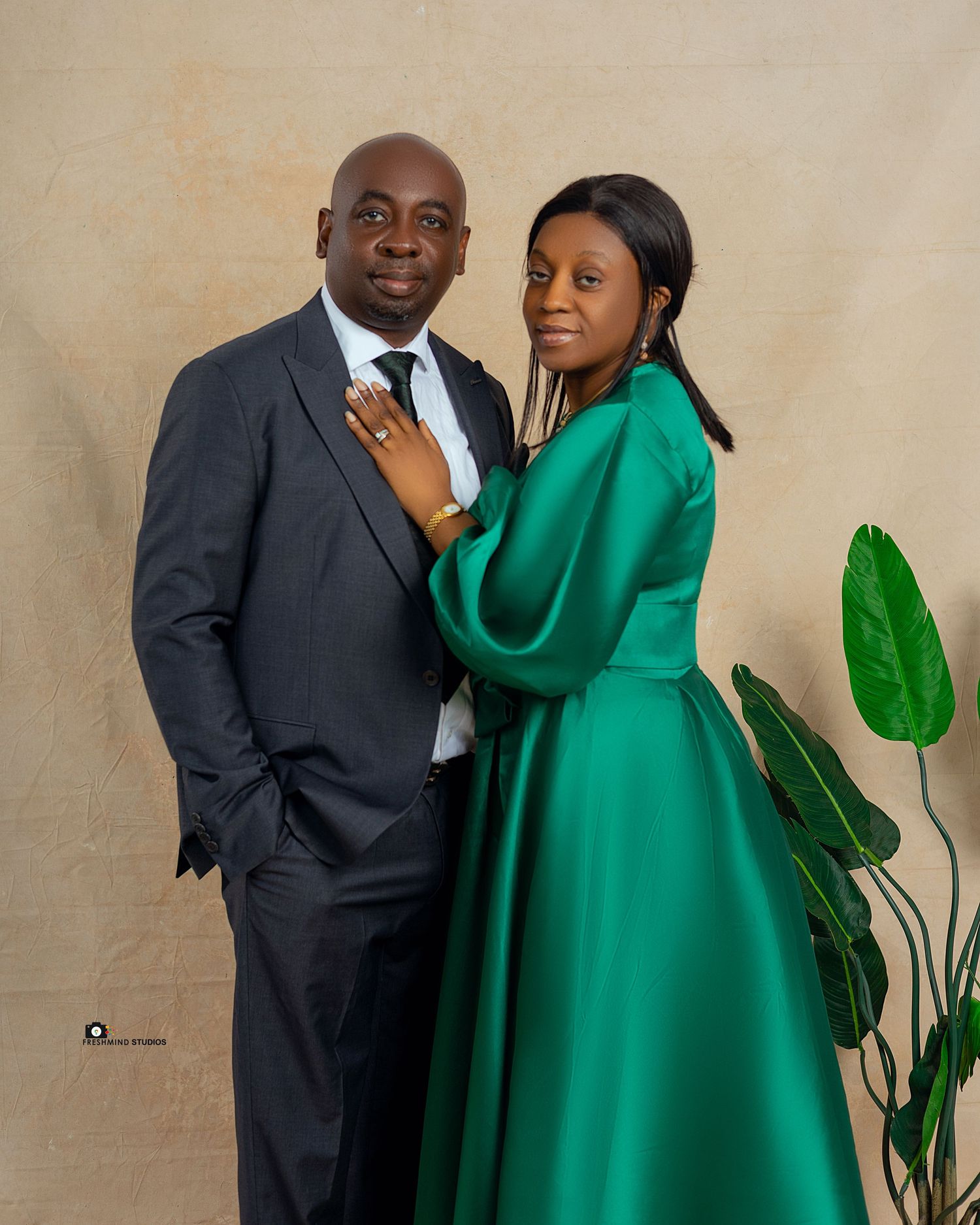 Elegant couple poses against neutral background with green foliage, wearing formal black suit and green dress.