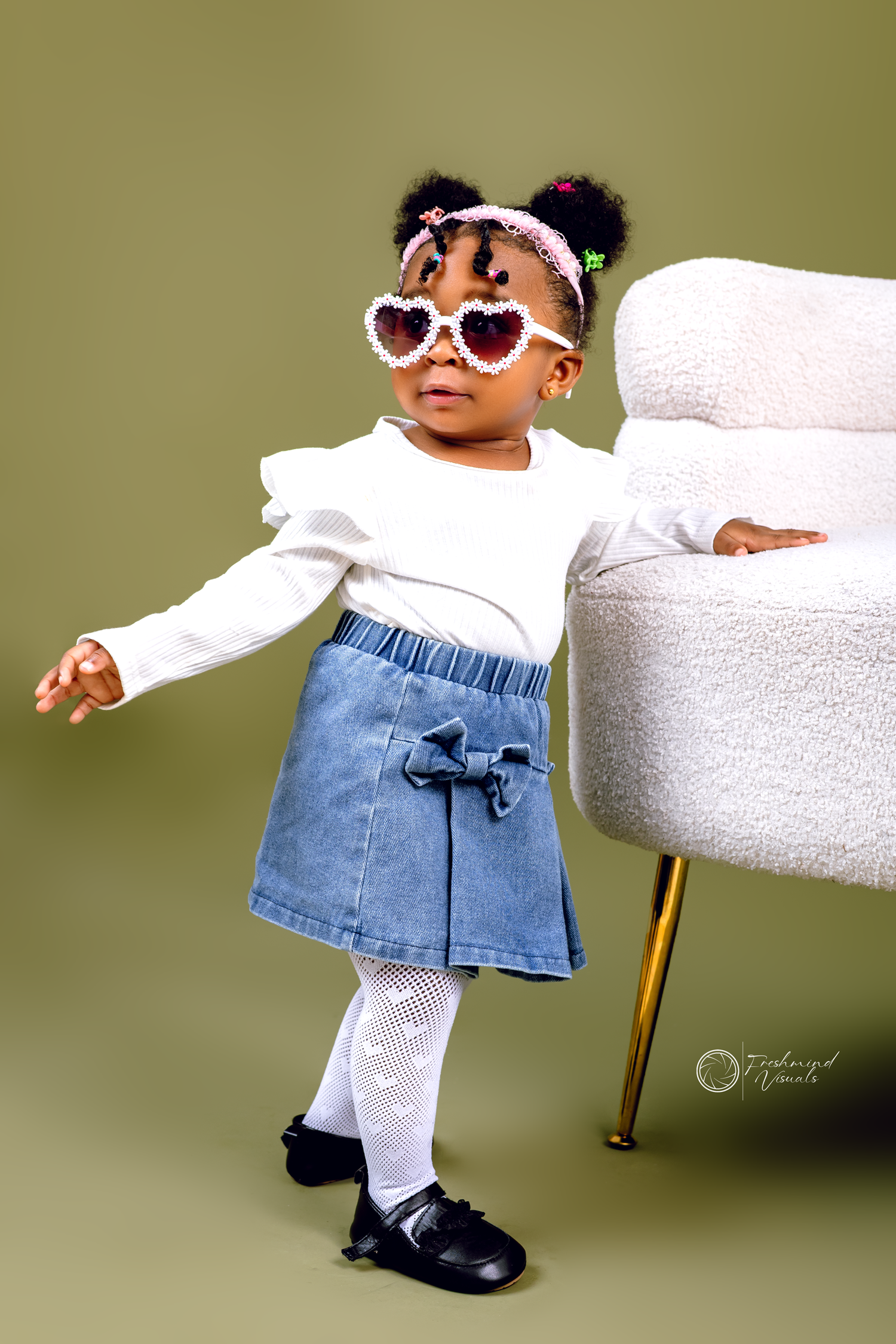 Toddler in white top and denim skirt posing playfully on white furniture.