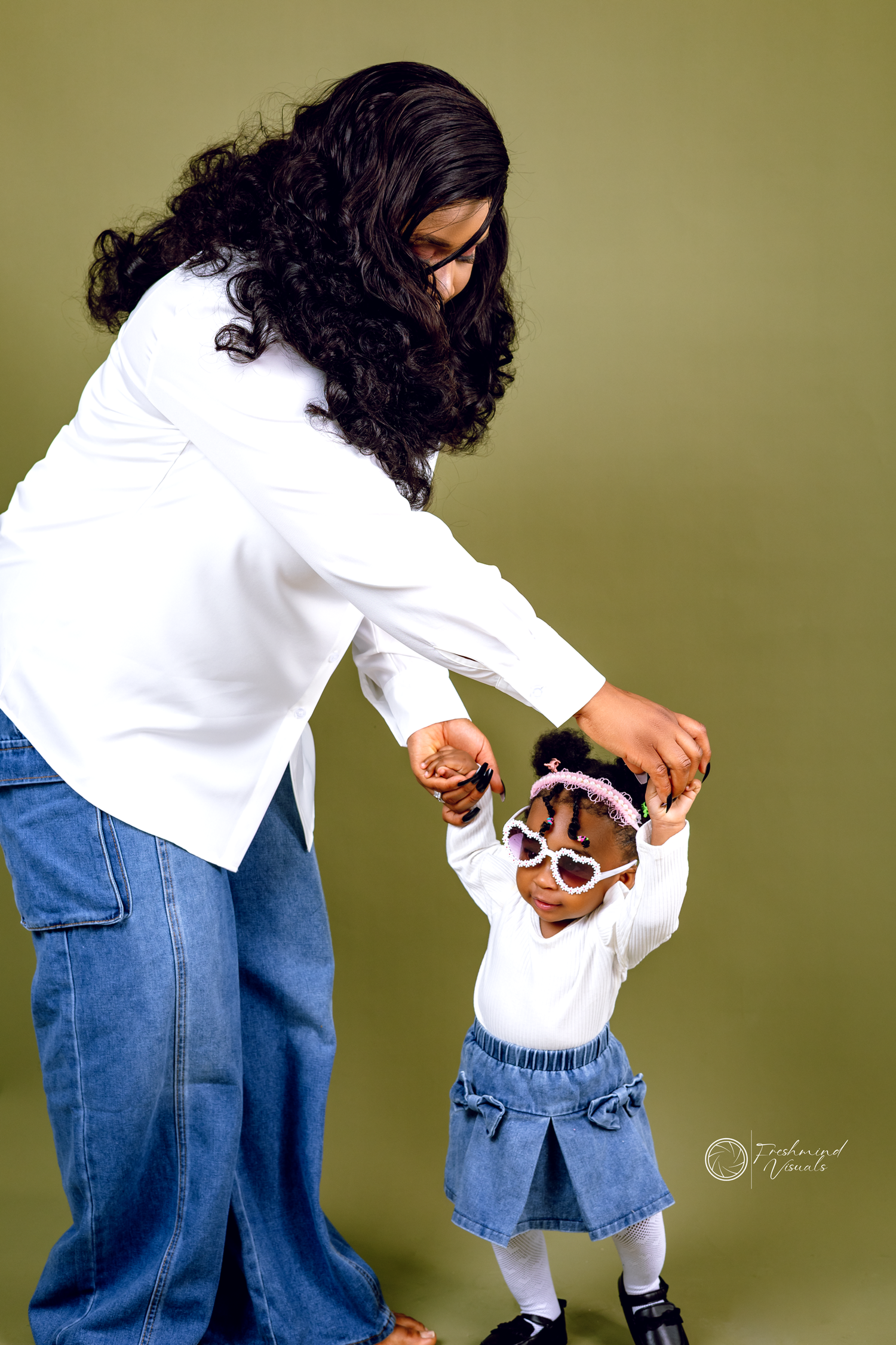 Person in casual white top and denim outfit poses playfully against olive green background.