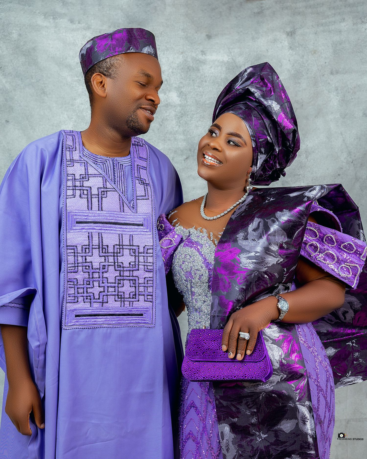 Couple in matching purple traditional African outfits posing against light backdrop.