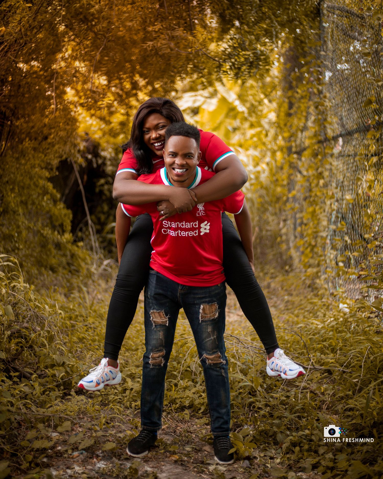 A playful couple in red shirts and jeans poses together in an autumn forest setting during a photo session.