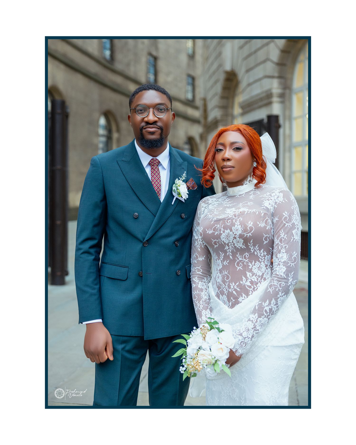 A couple poses together on their wedding day, with the bride in a lace gown and the groom in a teal suit.