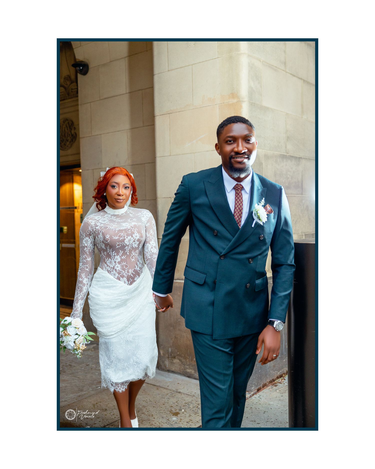 A couple in wedding attire walk together, with the bride in a lace dress and groom in a navy double-breasted suit.