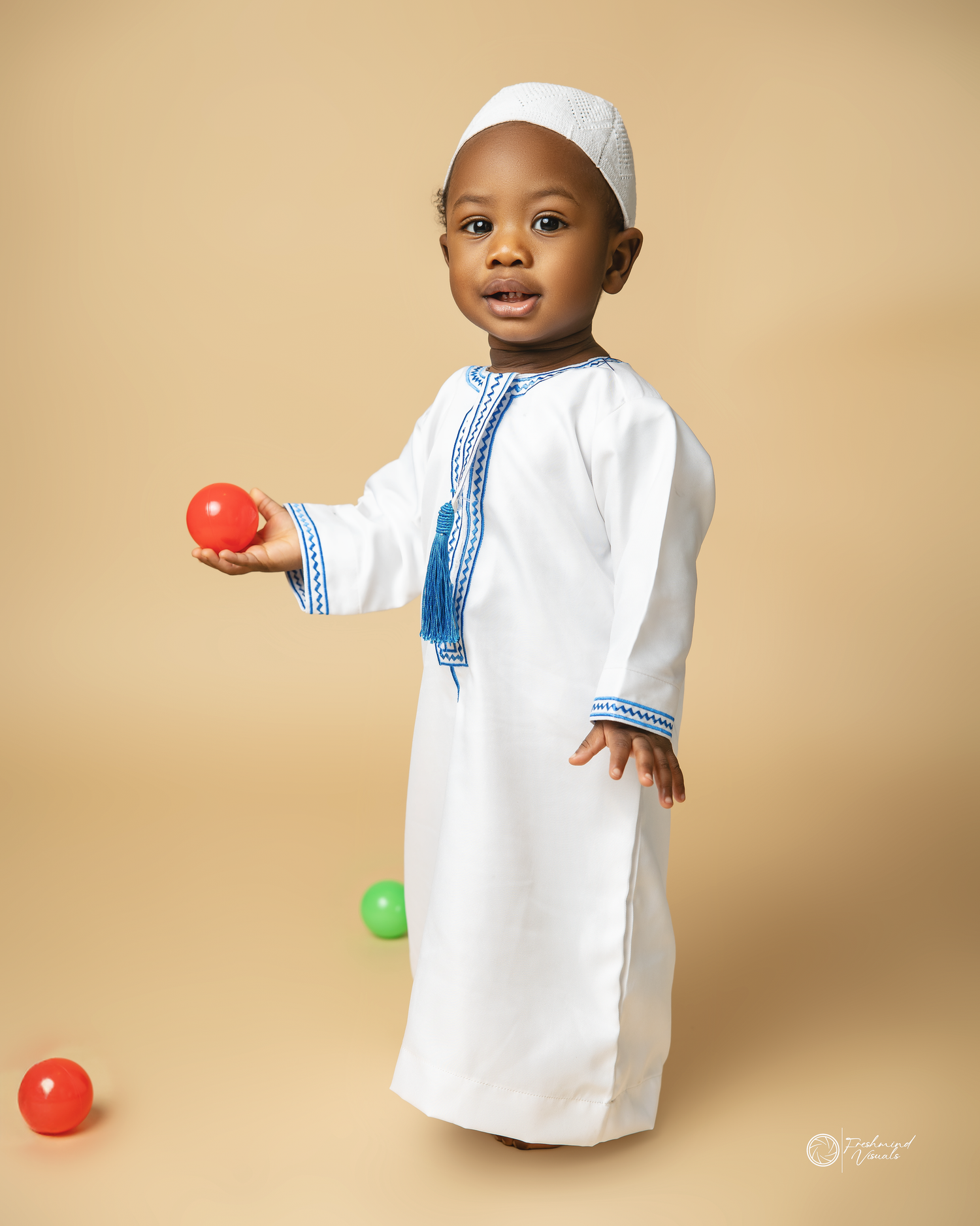 Toddler in white traditional Islamic dress holding colorful balls against beige background.