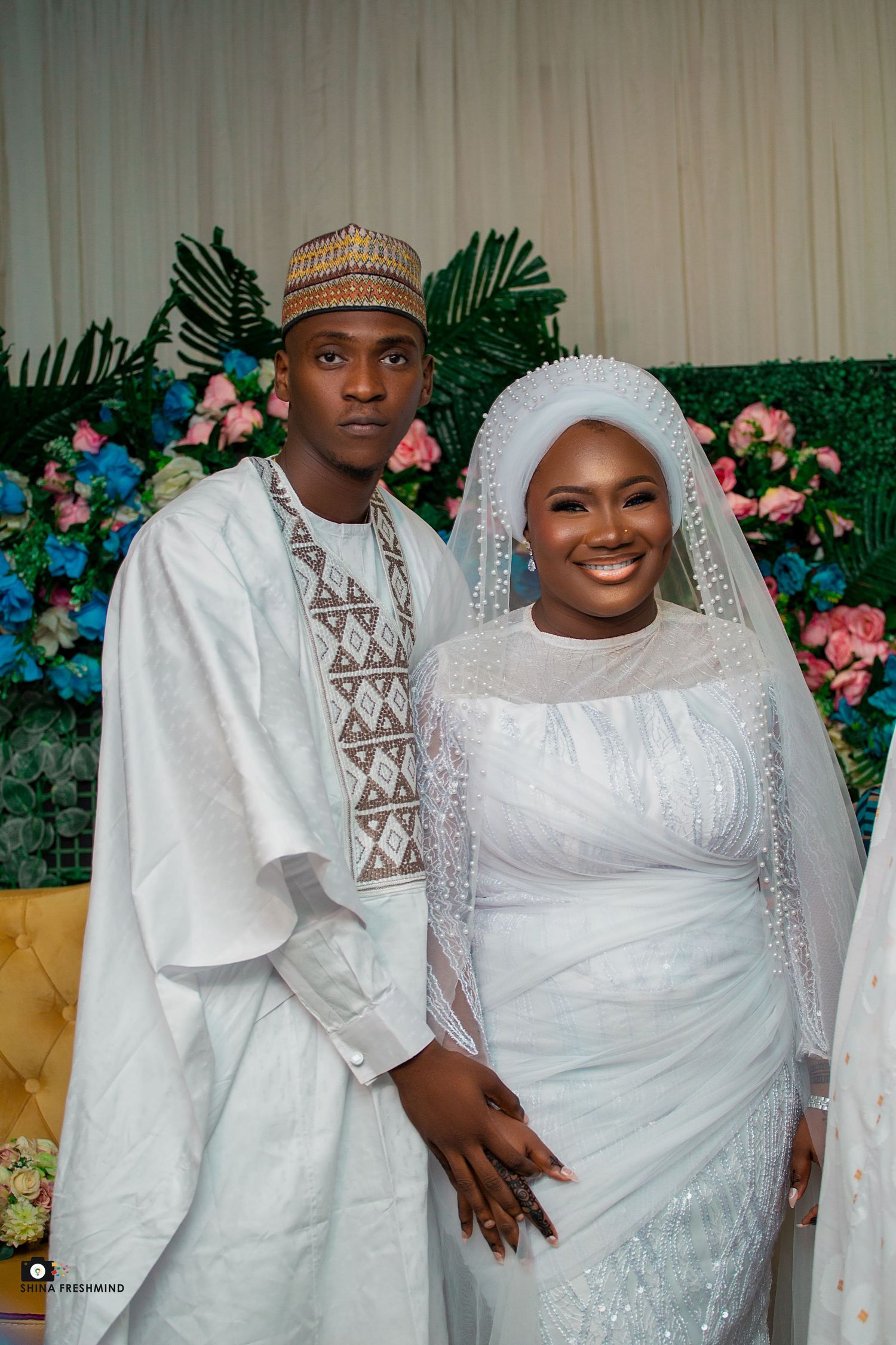Traditional wedding couple in matching white outfits with floral backdrop.